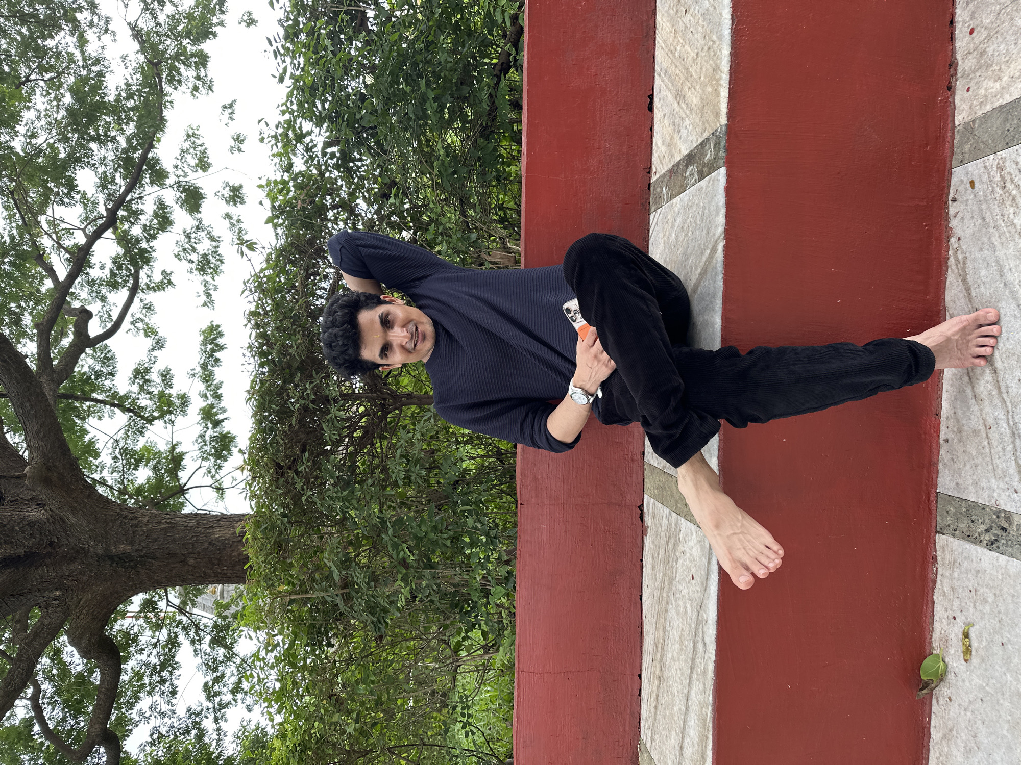 A man sits on a red bench with his left leg crossed over his right. He is wearing a dark blue long-sleeved shirt and black pants. His right arm is resting on his head, and he is holding a phone in his left hand. The bench is made of red painted concrete and has a marble seating area. The background consists of green bushes and a large tree. The ground is paved with light-colored tiles. The scene appears to be outdoors, possibly in a park or public garden in Shahapur, India.