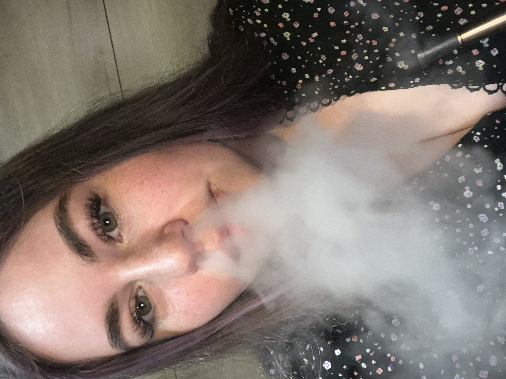 A young woman lies on a wood floor, exhaling a large cloud of vapor. She has fair skin, hazel eyes with long eyelashes, and dark brown hair with purple highlights. She wears a black dress with a small floral pattern. A metallic object is visible near her shoulder, possibly a vaping device. The location is unknown; there is no evidence to place it in Chaikovsky, Russia.