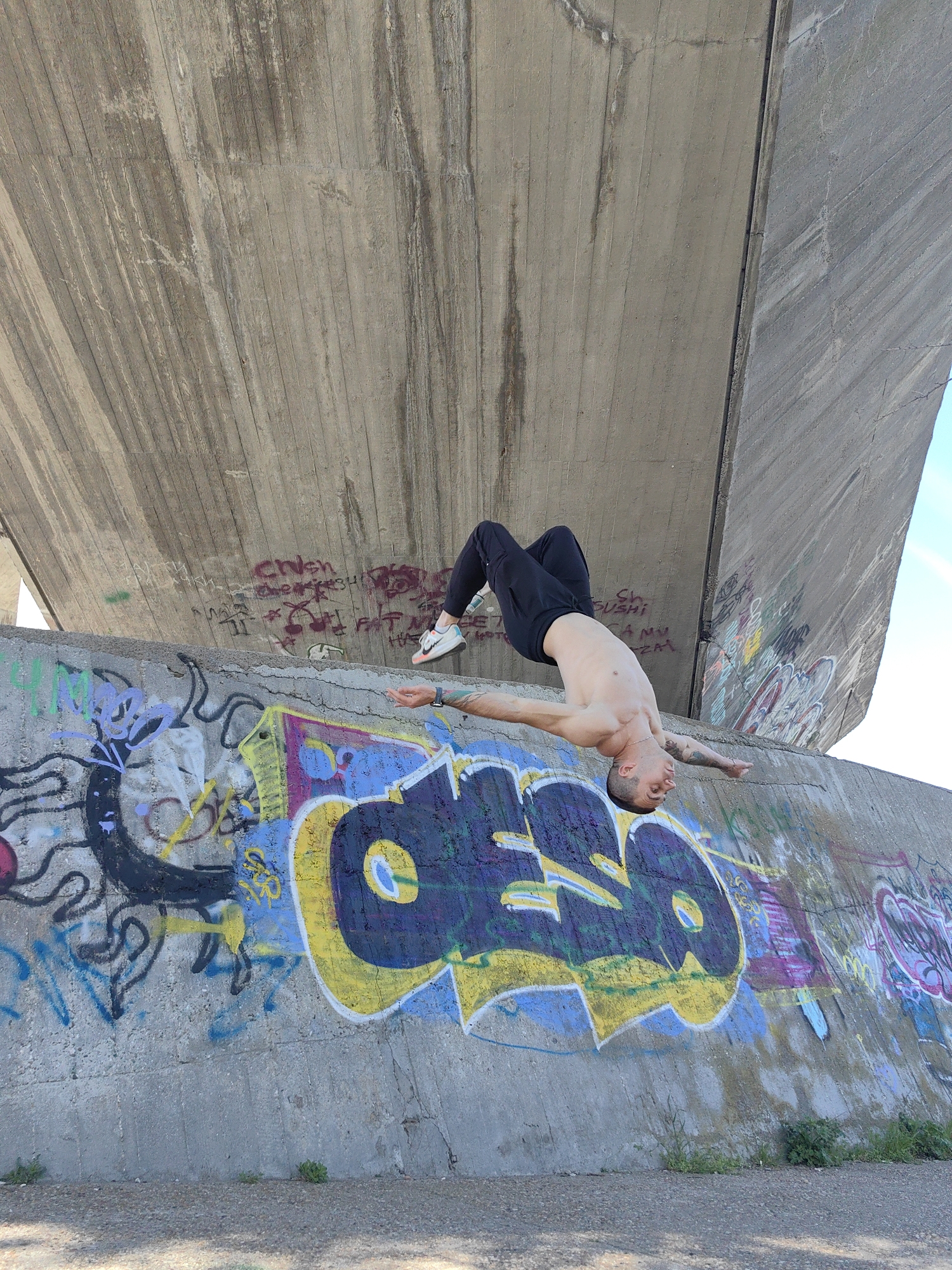 A man with short hair and tattoos is performing a backflip from the top of a concrete wall covered in graffiti. He is wearing black pants and white sneakers. The background consists of a large concrete structure, possibly a bridge support. The scene appears to be in an urban setting within the city of Varna, Bulgaria, suggested by the graffiti and architectural style. The weather seems sunny.