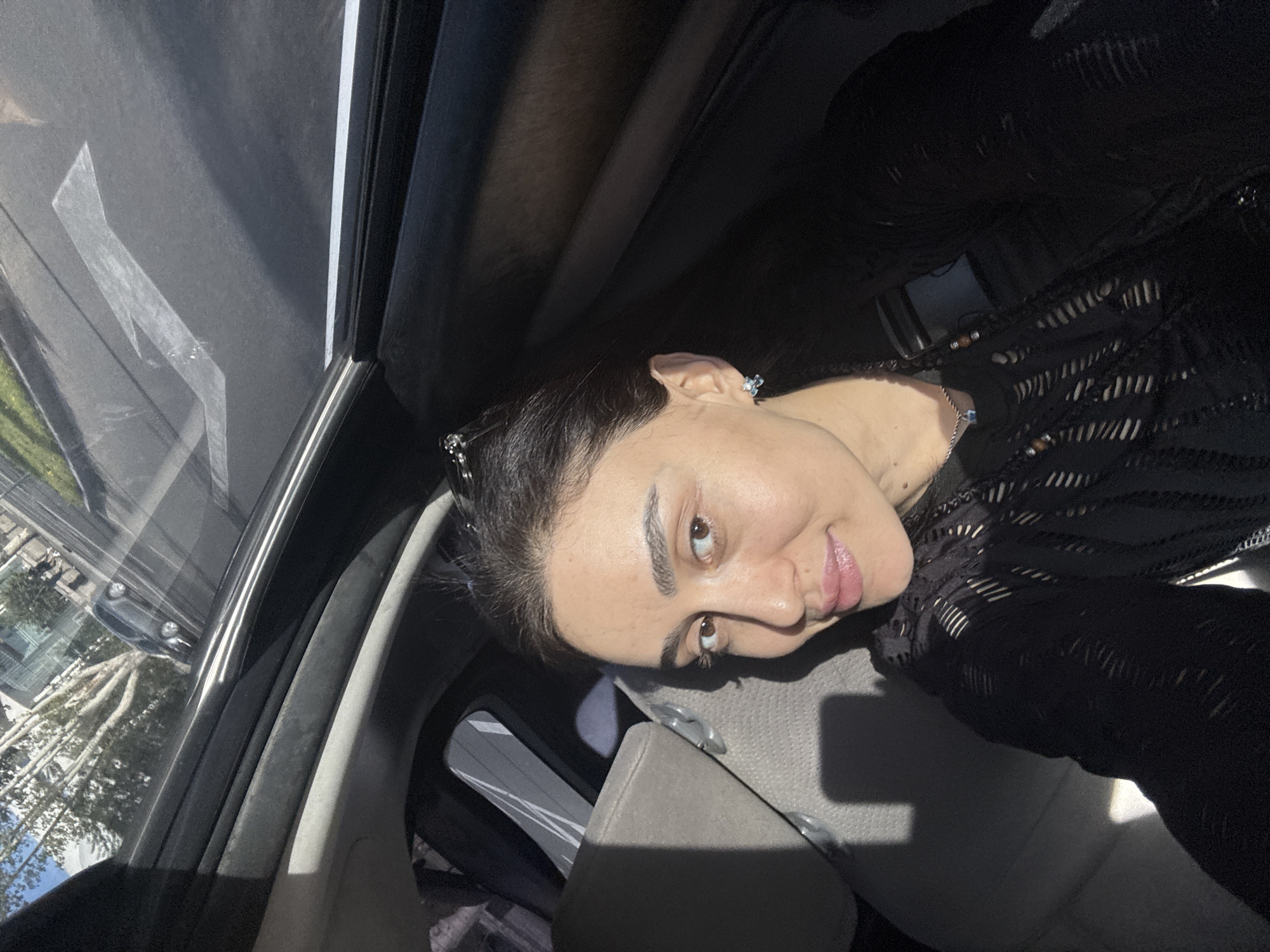 A woman is taking a selfie inside a car in Tbilisi, Georgia. The scene is illuminated by daylight. The woman has dark hair pulled back and is wearing a dark, patterned top. The car has a sunroof, and through the window, urban architecture and trees can be seen. Other cars are visible on the street. The woman is sitting in a light-colored car seat.