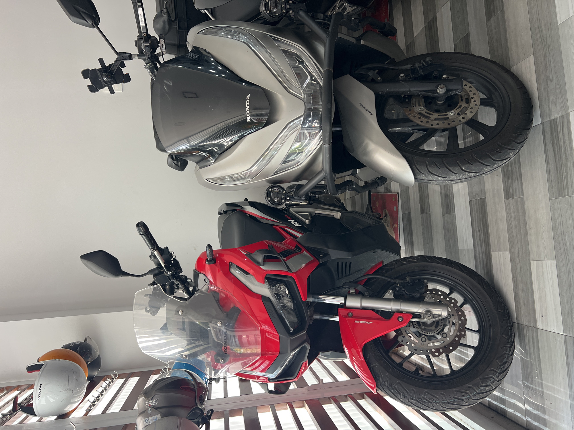 The image shows two Honda motorbikes, one silver and one red, parked inside what appears to be a garage or showroom. The silver motorbike is a Honda PCX. The red motorbike is a Honda ADV 150. There is a helmet rack in the lower left corner of the image. The location is possibly in Nha Trang, Vietnam.