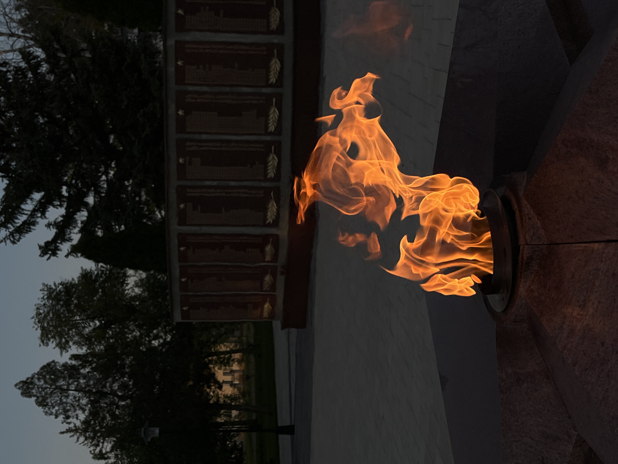 The image shows a burning eternal flame at a memorial in Novopokrovskaya, Russia. The flame is bright orange and dominates the right side of the image, coming from a metal structure. To the left of the flame is a stone structure with multiple plaques on it, each featuring a golden emblem and text. Behind the stone structure are trees, suggesting a park-like environment. The sky is dusky, indicating either dawn or dusk. The ground is paved with stone.