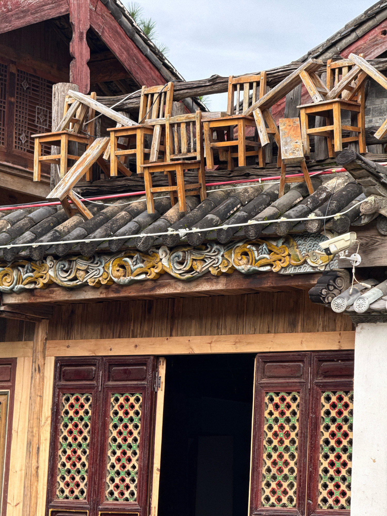 The image shows a collection of overturned wooden chairs on the tiled roof of a building in Yulong Nakhi Autonomous County, China. The chairs are stacked in a precarious manner, some on top of others, with their legs pointing upwards or sideways. The roof itself is covered with dark gray tiles, and a decorative wooden frieze runs along the eaves, featuring carved motifs in yellow and green. Below the roof, there is a wooden facade with a doorway revealing a dark interior. Above the roof, a red-painted wooden structure with a lattice window is visible. A security camera is mounted on the frieze, overlooking the scene. The sky is overcast. The context suggests a whimsical or artistic display rather than an accident, given the deliberate arrangement of the chairs.