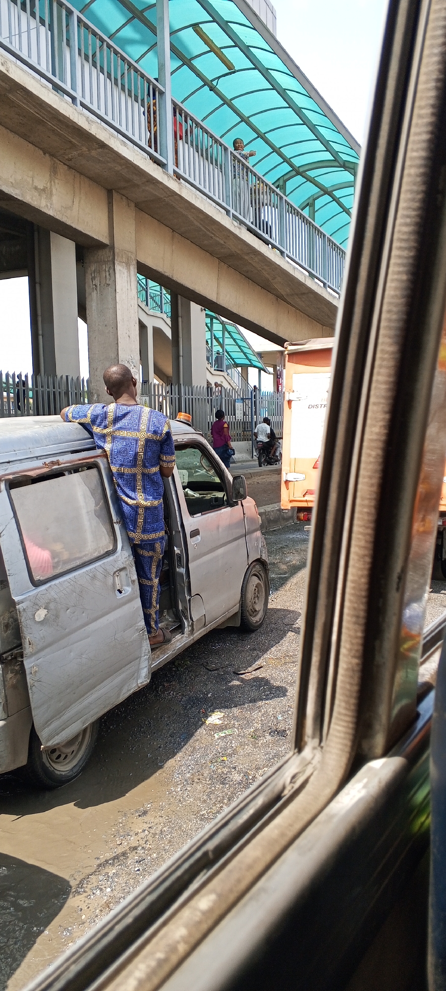 In Agbani, Nigeria, a person wearing a blue and yellow patterned garment stands with their back to the camera, leaning on the open door of a silver van. The van appears worn, with visible scratches and dents on its exterior. The person's feet are positioned on the ground, and they are looking upwards towards an elevated pedestrian walkway.
The walkway, visible in the background, has a translucent turquoise roof supported by gray metal beams and railings. Several other people are present on the walkway, some appearing to be engaged in conversation. Below the walkway, a fence separates it from the street.
On the street level, the van is parked on a surface that appears to be a mix of gravel and dirt, with some muddy patches. Further down the street, an orange truck is visible. The overall scene suggests an urban environment with ongoing activity, potentially during daylight hours given the bright lighting and shadows.