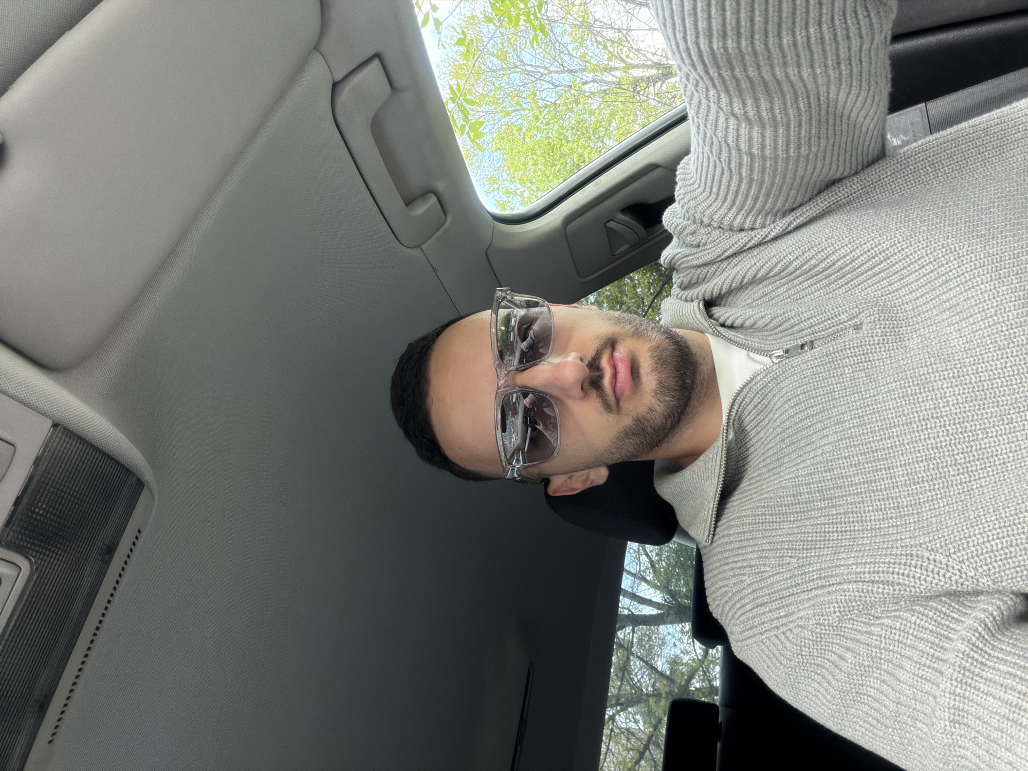 A vertical, low-angle shot captures a young man inside a car. He is centered in the frame, looking directly upwards towards the camera, suggesting a selfie or a photo taken from a low vantage point. The man wears clear-framed sunglasses, which reflect a distorted view of the car's interior. He has short dark hair, a trimmed beard or stubble, and his lips are slightly pursed. He is dressed in a light grey, ribbed knit sweater featuring a visible zipper at the collar.
The setting is the interior of a vehicle, with the light grey fabric of the car's roof lining prominent. Above the man, a sunroof offers a clear view of the outside world, revealing bright green tree canopies against a light blue or bright sky, indicating it is daytime with good weather. Parts of the car's overhead console and a sun visor are also visible, reinforcing the indoor-car setting. The focus is primarily on the man and the immediate environment inside and just above the car. No specific activity is depicted other than posing for the photograph. There is no visible text. The location is Plovdiv, Bulgaria.