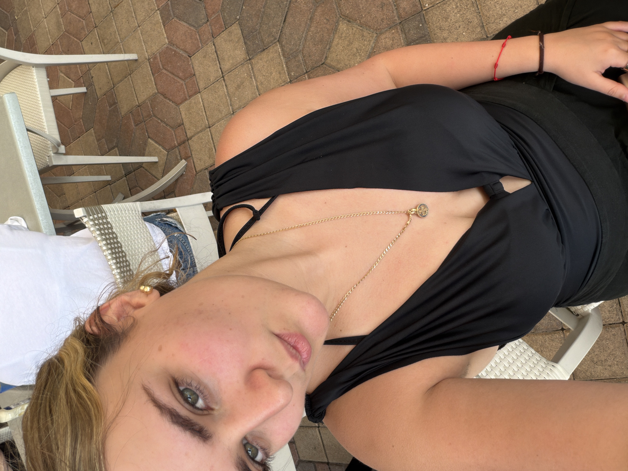 A woman is sitting outside on a patio in Key Biscayne, United States of America. She is wearing a black top and a gold necklace with a charm. She has blonde hair and green eyes. She is also wearing two bracelets on her right wrist. A white and gray chair is visible to the left of her. The patio is paved with red and brown bricks.
