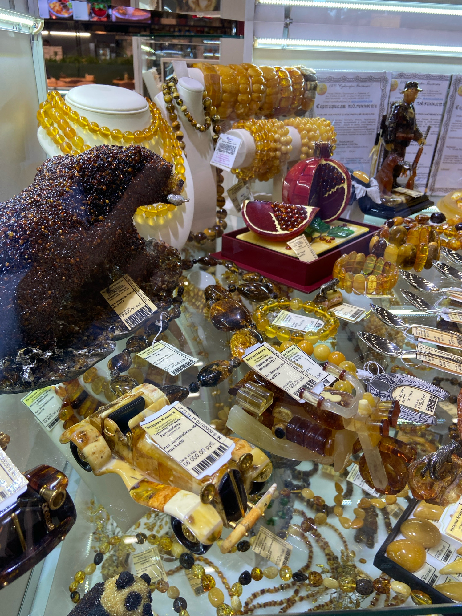 The image shows a display case filled with amber jewelry and carved objects. Prominently featured are necklaces, bracelets, a car sculpture, a panda sculpture, and what appears to be a pomegranate sculpture. Several items have price tags attached, with prices indicated in Russian Rubles. In the background, there are display boards with text in Russian and a small figurine of a soldier with a dog. The overall environment suggests a retail store specializing in amber products. There are no people visible in the image. The city of Ob, Russia, is not directly identifiable from the visual cues in the image; this information is likely external context.
