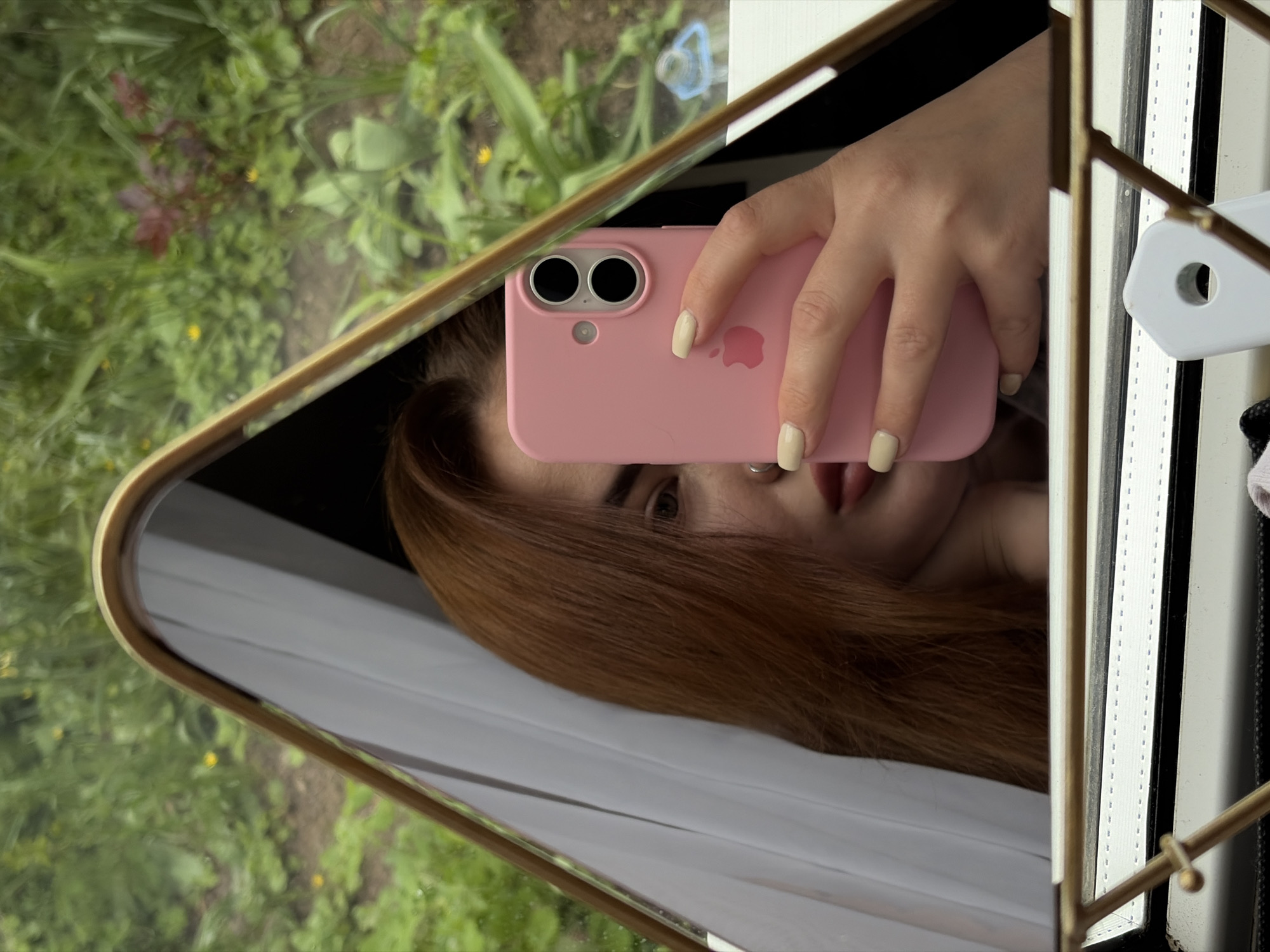 The image shows a person taking a selfie in a mirror. The person is holding a pink iPhone, and their face is partially visible. They have light skin, brown hair, and are wearing a nose ring. The mirror has a gold frame and is positioned in front of a window that overlooks a green outdoor area. The reflection in the mirror also shows a white curtain and part of the mirror's stand. The location is assumed to be Novopokrovskaya, Russia.