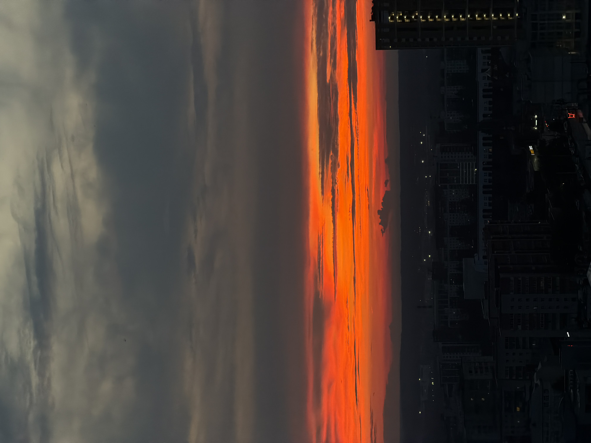 The image captures a city skyline during a vibrant sunset. The sky is dominated by a fiery orange and red gradient, transitioning to dark gray clouds at the top. Below the dramatic sky, a cityscape is visible, shrouded in the dim light of dusk. Numerous buildings, varying in height and architectural style, are illuminated by scattered lights. A tall, modern building with vertical lines of illuminated windows stands prominently on the right side of the frame, suggesting an urban environment. The overall impression is one of a metropolitan area at the end of the day, bathed in the dramatic colors of a sunset. The location appears to be the city of Perm, Russia.