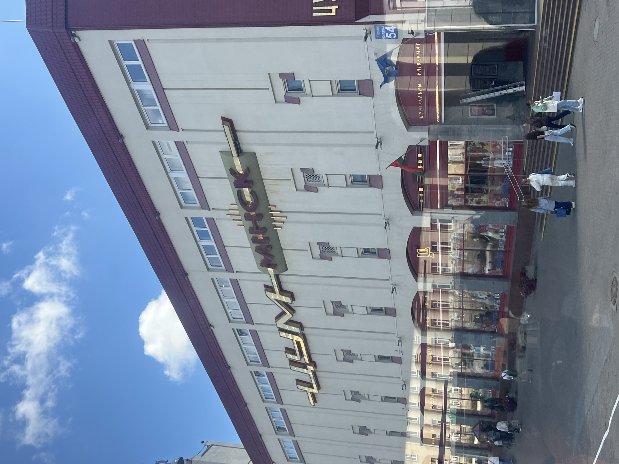 The image shows the TsUM (Central Universal Department Store) in Minsk, Belarus, on a sunny day. The building is a large, multi-story structure with a distinctive maroon roof and cream-colored facade. Large, golden Cyrillic lettering on the building spells out "ЦУМ МИНСК" (TSUM MINSK). Below the main lettering, there are several arched entrances with large glass windows, showcasing mannequins and merchandise inside. People are visible walking on the sidewalk in front of the store, some carrying shopping bags. Two flags, one red and green and another blue, are displayed outside one of the entrances, along with a sign indicating a street number "54". The sky is blue with scattered white clouds. The overall scene depicts a busy urban commercial area.