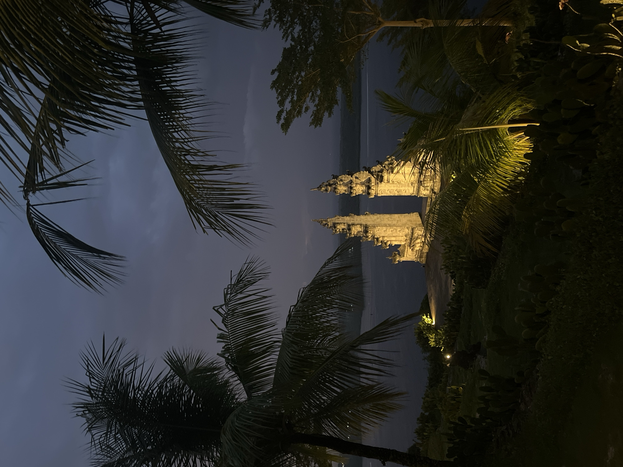 This outdoor shot, taken at dusk or night, depicts the Pura Batu Medau temple complex in Nusa Penida, Indonesia. The temple is lit up with golden lights and stands between palm trees and other lush vegetation. The sky above is dark blue, indicating nighttime. The scene offers a glimpse into the serene ambiance surrounding this temple, set against the backdrop of the natural landscape.