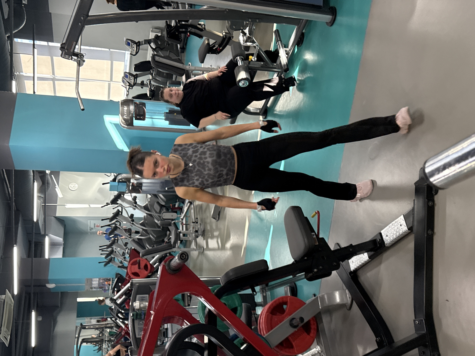 The image depicts an indoor scene at a gym. A young woman stands in the foreground wearing a gray leopard-print tank top and black leggings. She is stretching, with her arms outstretched. Another woman is visible in the background, seated on exercise equipment, wearing all black. The gym contains a variety of exercise machines and equipment. The location is Yekaterinburg, Russia, based on visual characteristics potentially related to the gym design and equipment present.