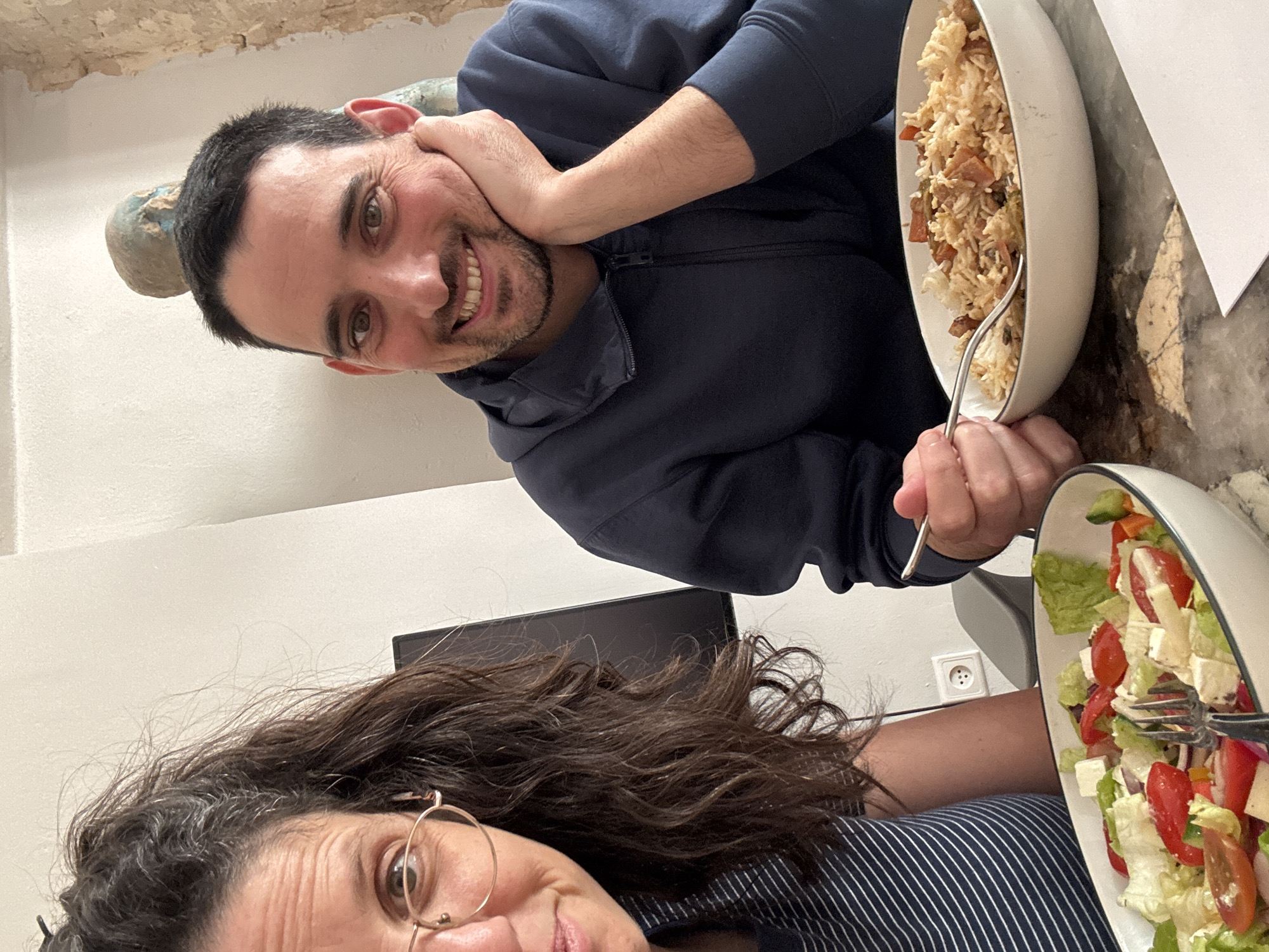A selfie-style photograph, captured at a 90-degree clockwise rotation, shows two individuals sharing a meal indoors, likely in Tel-Aviv, Israel.
In the foreground, partially visible on the left, is a woman with long, dark, wavy hair and round, gold-rimmed glasses. Only her right eye, forehead, and part of her cheek and mouth are visible as she looks towards the camera, presumably taking the picture. She wears a striped top. Resting on the table next to her, a white oval bowl contains a fresh salad with green lettuce, red tomatoes, red onions, and cubes of white feta cheese, with a fork inside.
Behind and to the right of the woman is a man with short dark hair and a beard, wearing a dark blue hooded sweatshirt. He smiles broadly at the camera, his eyes wide and engaging. His left hand is casually placed near his head, while his right hand holds a fork in a white oval bowl filled with rice, mixed vegetables, and what appears to be nuts or other solid ingredients.
The table surface is a light-colored, possibly marble or stone pattern. A white piece of paper lies on the table near the rice bowl. The background consists of a plain light-colored wall, with some textured material near the ceiling. A dark rectangular object, possibly a tablet, is visible behind the woman's head, and a white electrical outlet is present on the wall behind the man's arm. The cheerful expression of the man suggests a pleasant and casual atmosphere during the meal. No visible text is present.