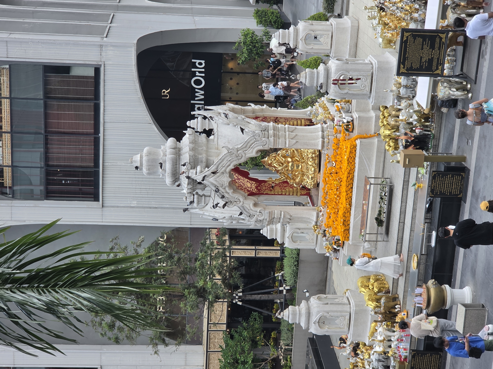 The image shows the Erawan Shrine, a popular Hindu shrine in Bangkok, Thailand. The shrine, located near the Grand Hyatt Erawan Hotel, is dedicated to Brahma, the Hindu god of creation. It's adorned with golden figures, garlands, and surrounded by offerings. People are praying and making offerings at the shrine. The background shows part of the cityscape with the CentralWorld shopping mall visible.