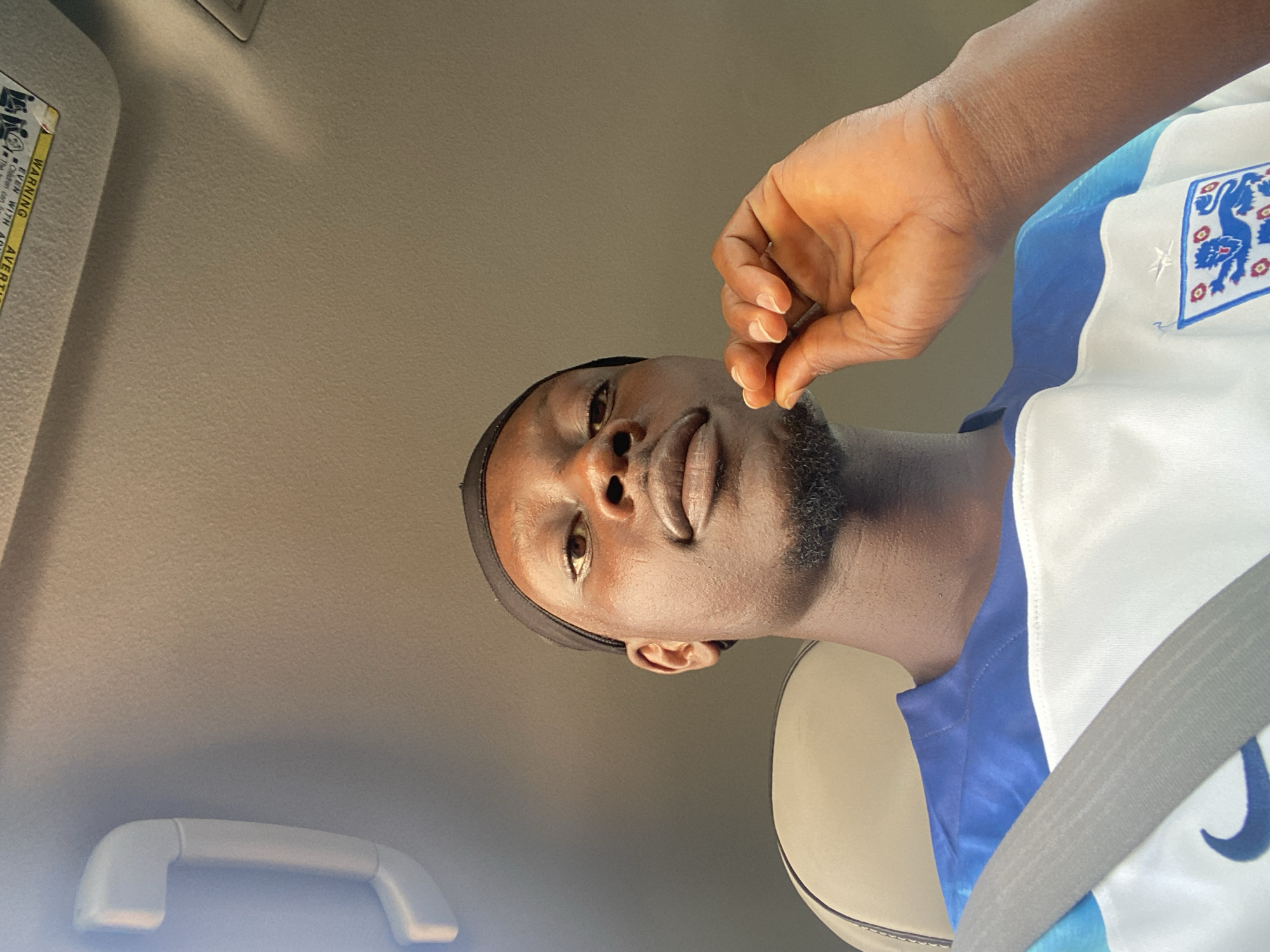 A man is sitting in the passenger seat of a car, looking at the camera. He is wearing a black headscarf and a white and blue jersey with a crest on the chest. His right hand is raised to his chin. The interior of the car is visible, with a beige seat and a grey seatbelt. The ceiling of the car, which has a warning label, is also visible. The image appears to have been taken outdoors, as suggested by the lighting. The background is out of focus, suggesting a shallow depth of field. It is not possible to determine the specific location within Bar Jirgi Summa, Nigeria from the image alone.