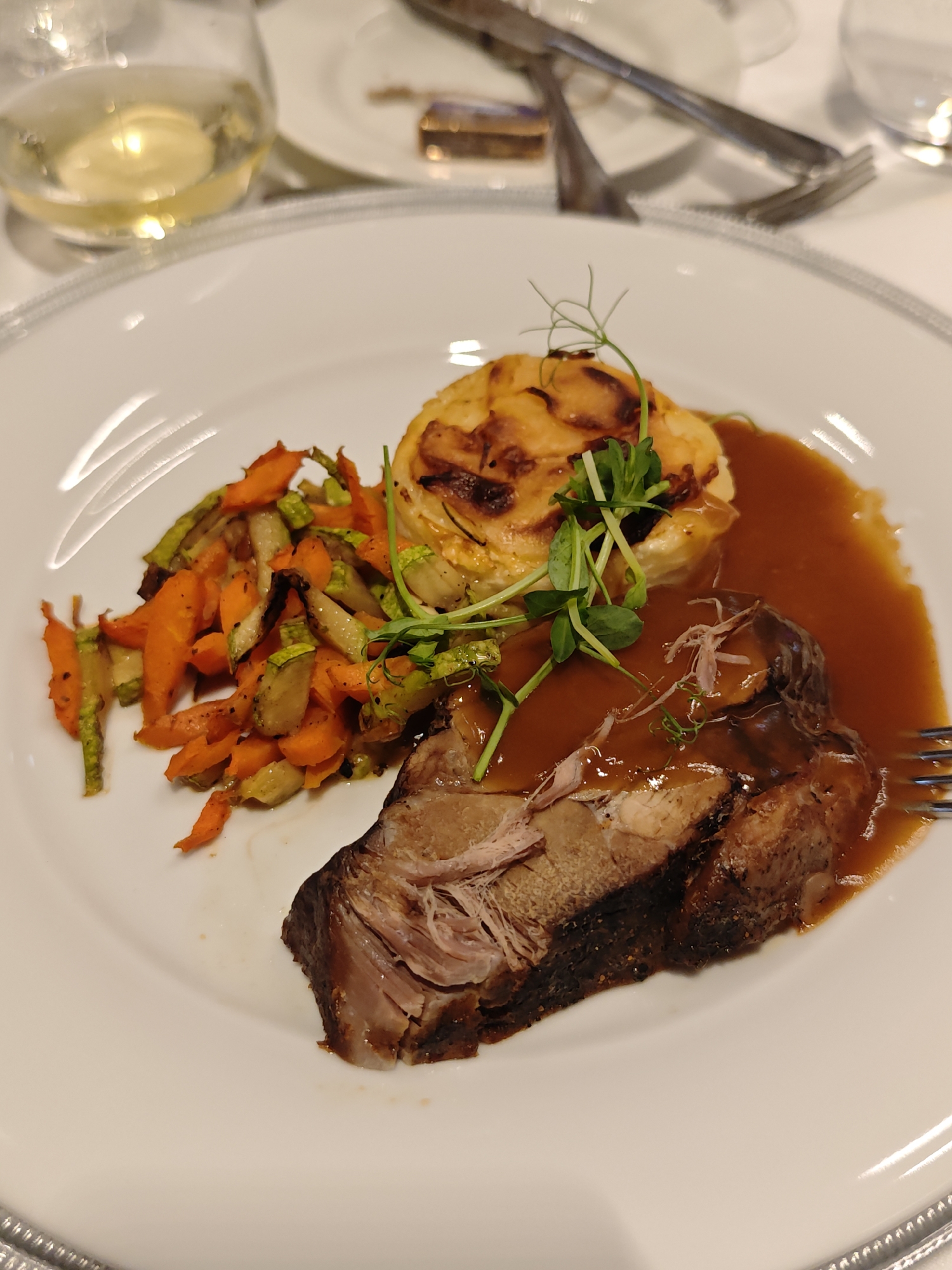 A close-up photograph shows a white plate featuring a meal. The plate is edged with a silver border. The meal consists of a slice of meat covered in gravy, a potato gratin, and cooked carrots and zucchini. Fresh sprouts are placed atop the potato gratin. A fork is visible on the right side of the plate, partially obscured by the meat. In the background, a wine glass filled with a light-colored liquid sits atop a white table. A set of silverware rests on a white surface, further back in the image. The photograph appears to have been taken indoors. There is no evidence to place this photo in Sofia, Bulgaria.