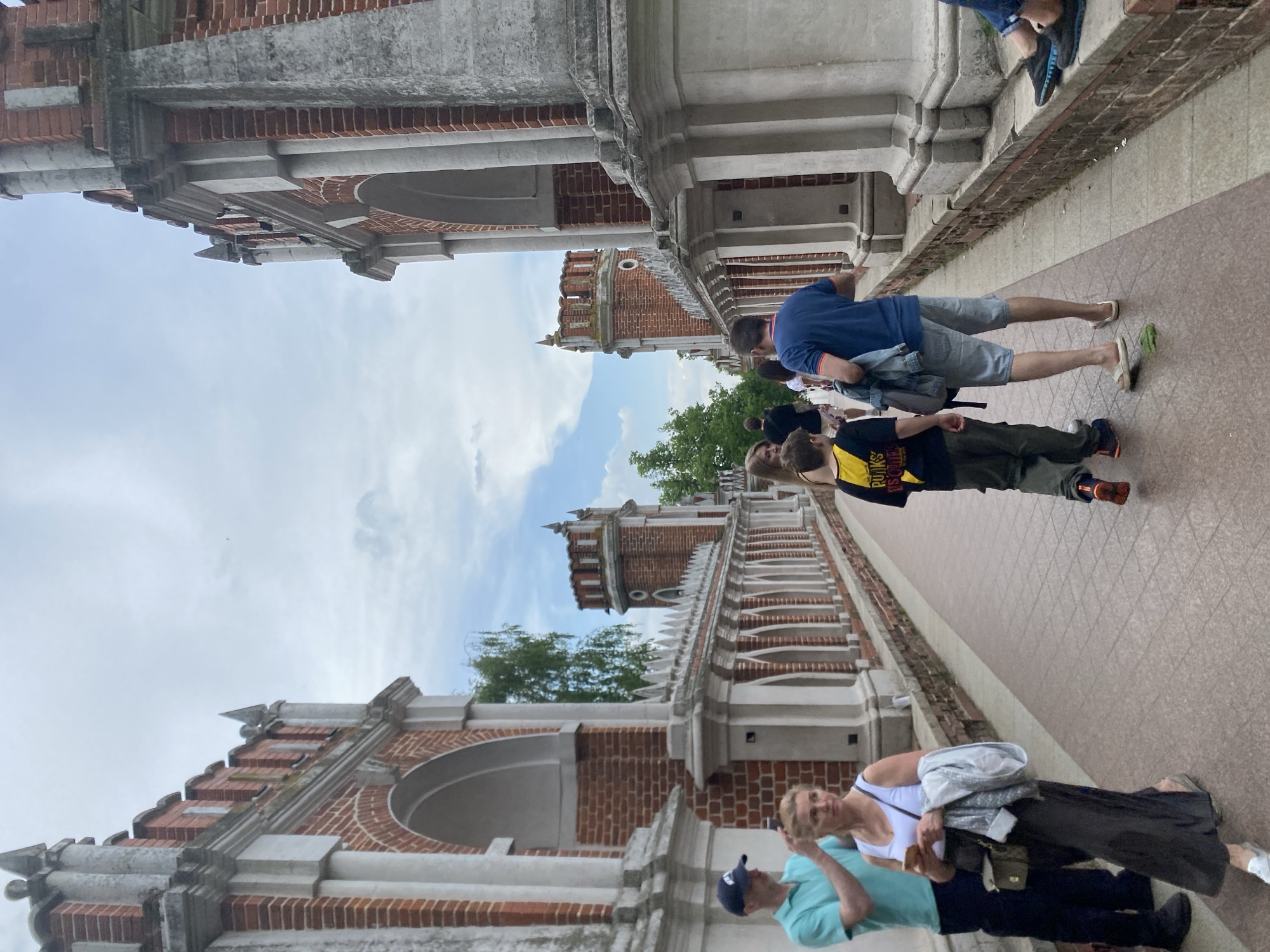 A low-angle, medium shot shows a group of tourists walking along a paved walkway lined with brick and white-trimmed buildings under a partially cloudy sky. The image orientation is unusual, tilting the scene so that the walkway appears to be angled upwards from left to right.
Four people are visible: two men, a woman, and a child or young adult. On the right side, a man wearing a blue shirt and gray shorts walks alongside a child in a yellow and black shirt and olive pants. On the opposite side, a woman in a white shirt and dark skirt is accompanied by a man wearing a light blue shirt and dark baseball cap. They are walking in the same direction as the first pair.
The buildings have a distinctive architectural style featuring red brick and white stone trim. The arched windows and decorative elements suggest historical architecture. The background includes green trees and a cloudy sky with patches of blue. The location is Unknown.