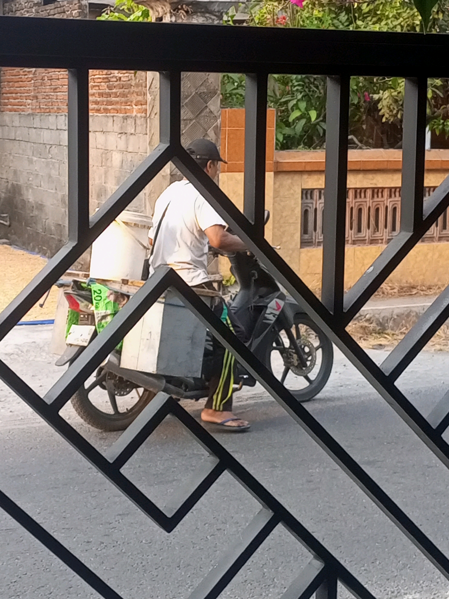 In Ngasem, Indonesia, a man wearing a black cap and a white t-shirt rides a motorcycle. He is seated on the motorcycle, which has a gray storage box mounted on the back. A white bucket is also attached to the rear of the motorcycle. The man is wearing striped pants and flip-flops. The motorcycle is on an asphalt road. In the background, there is a brick wall and some foliage. The scene is framed by a black metal fence with a diamond pattern.