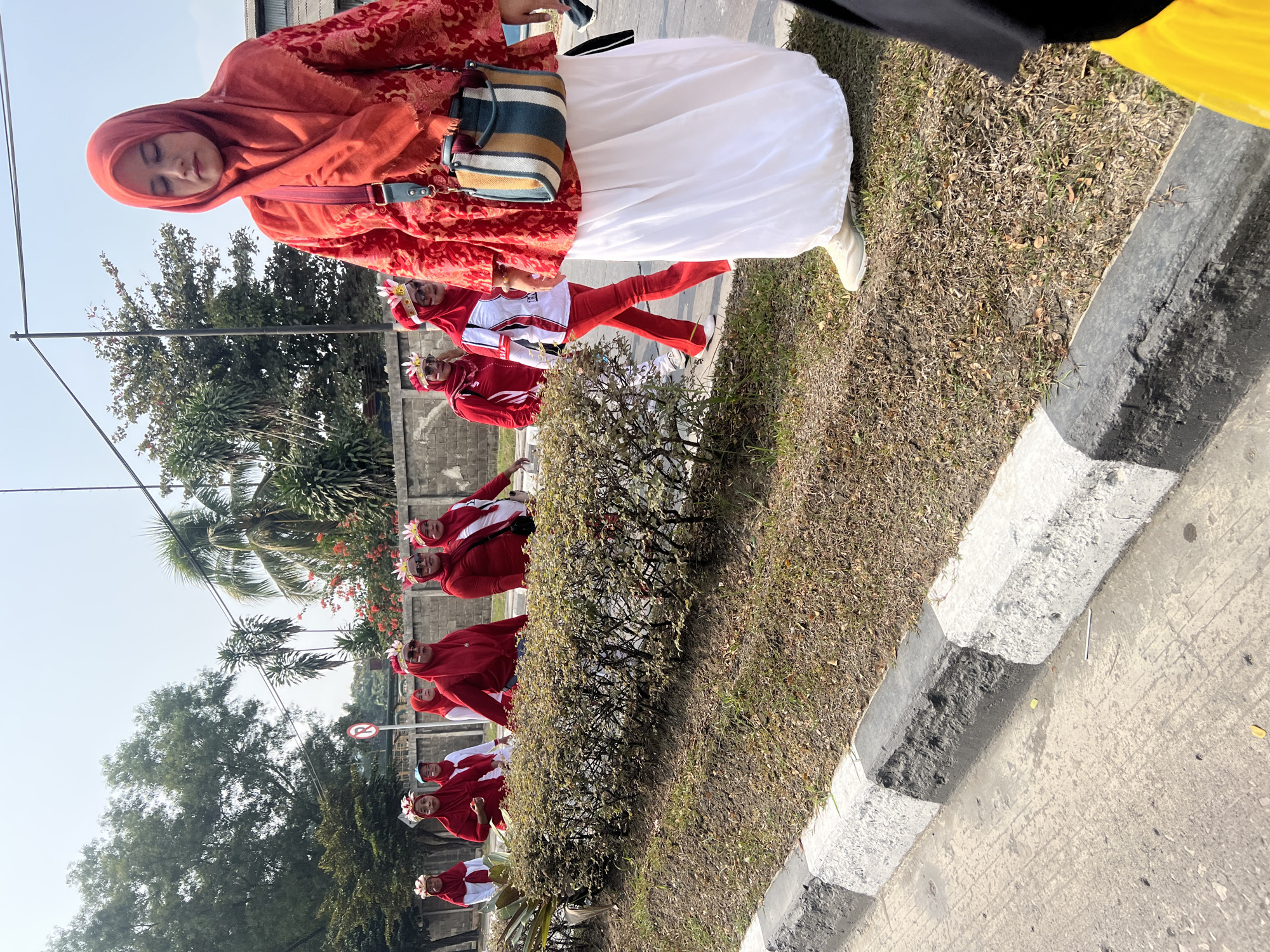 In Klapanunggal, Indonesia, a group of women wearing red and white attire and headdresses are walking along a paved road. The woman in the foreground, in a bright red patterned tunic and a long white skirt, carries a striped cross-body bag. Behind her, several other women are walking in a procession, also in red and white outfits and floral headdresses. They are walking past a low, manicured hedge and a gray and white striped curb. Trees and a concrete wall line the background. The scene appears to be a parade or cultural event.