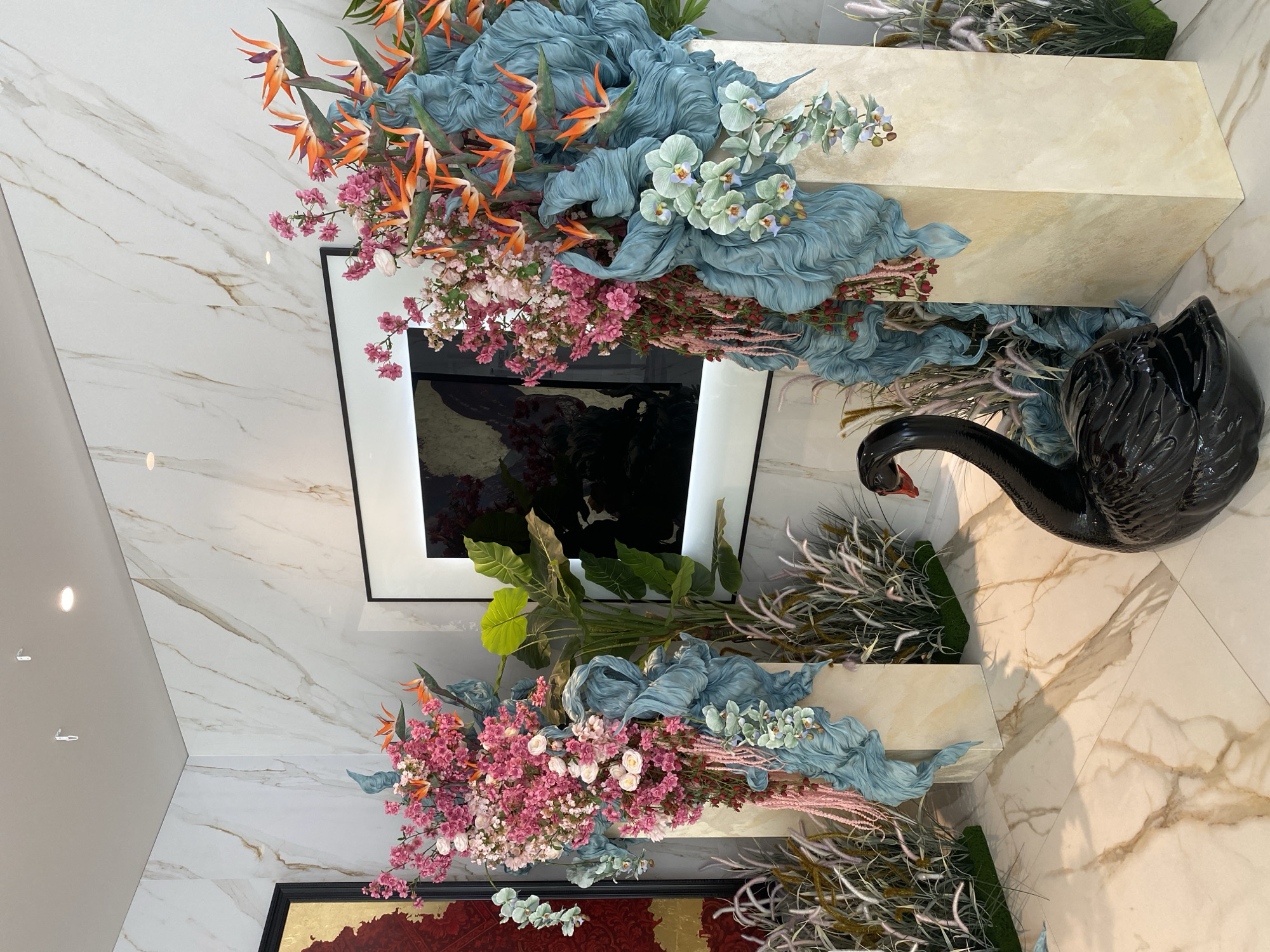 The image shows an interior decoration, possibly in a luxury setting. Two rectangular marble pedestals display floral arrangements featuring pink blossoms, Birds of Paradise flowers, and blue fabric resembling draped water. A black swan statue is placed near one of the pedestals. In the background are framed artworks on a white marble wall, one with a black background. The environment appears to be a modern, well-lit space with marble flooring and wall paneling. No people are visible.
The city of Sochi, Russia is not clearly referenced or identifiable from the provided image. There are no identifying landmarks or features to verify if the image originates from that location.