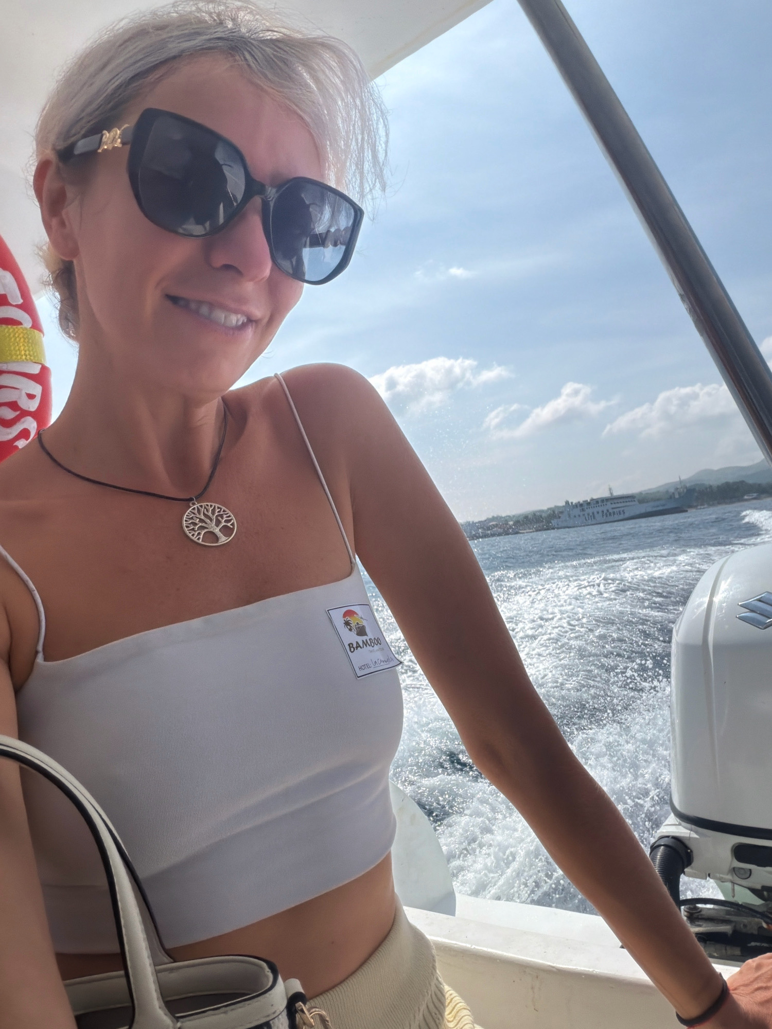 A woman is taking a selfie on a boat in the waters of Malay, Philippines. She is wearing sunglasses, a white crop top, and a necklace with a tree of life pendant. She has a name tag on her top that reads "BAMBOO HOTEL LA CARMELA". In the background, there is a ferry boat and a mountainous coastline. The water is choppy, creating a spray that is visible around the boat. The sky is blue with some white clouds.