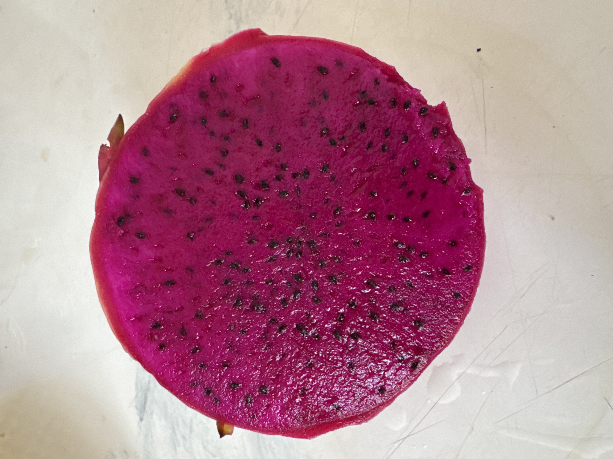 The image shows a cross-section of a vibrant pink dragon fruit with numerous small black seeds embedded within the pulp. The fruit appears freshly cut, with a slightly uneven edge suggesting it was likely cut by hand. A small portion of the outer skin, showing remnants of green, tan, and pink hues, is visible on one side. The fruit rests on a smooth, light-colored surface that may be a countertop or plate. There are no people or other objects visible in the immediate scene. The location in Kuta Selatan, Indonesia, is not discernible from the image.