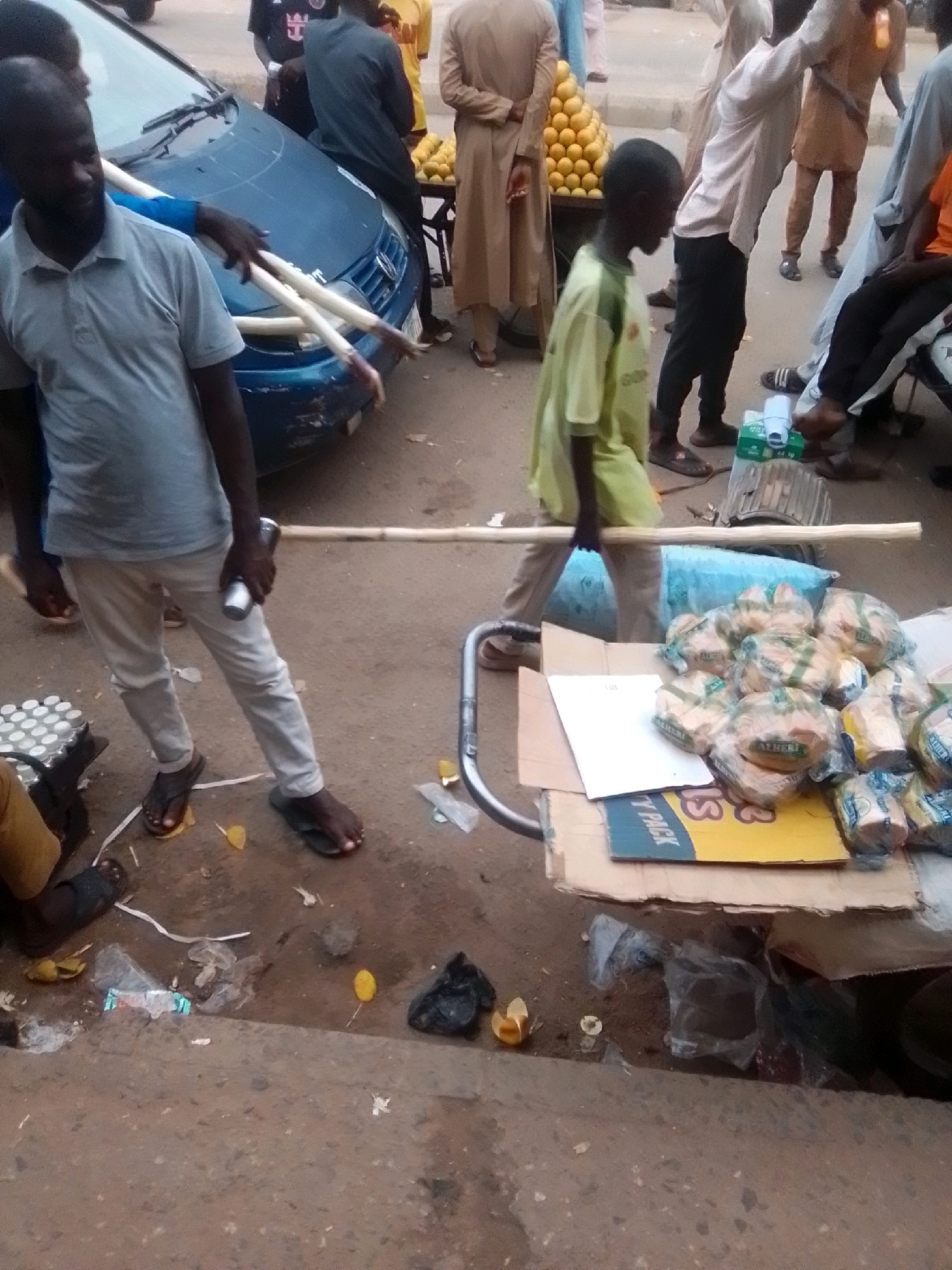 In the city of Arawa, Nigeria, a street scene unfolds. A man, wearing a light grey polo shirt and white pants, stands next to a blue car, holding a long wooden stick. He is accompanied by another person whose arm, wearing a blue sleeve, is near the stick. In the background, a vendor pushes a cart laden with bread loaves wrapped in plastic. The cart also carries a stack of what appear to be canned drinks. Further in the background, a pile of oranges is visible on a stand, and several people, some in traditional attire, are present. The ground is littered with debris, including plastic bags, wrappers, and fallen fruit.