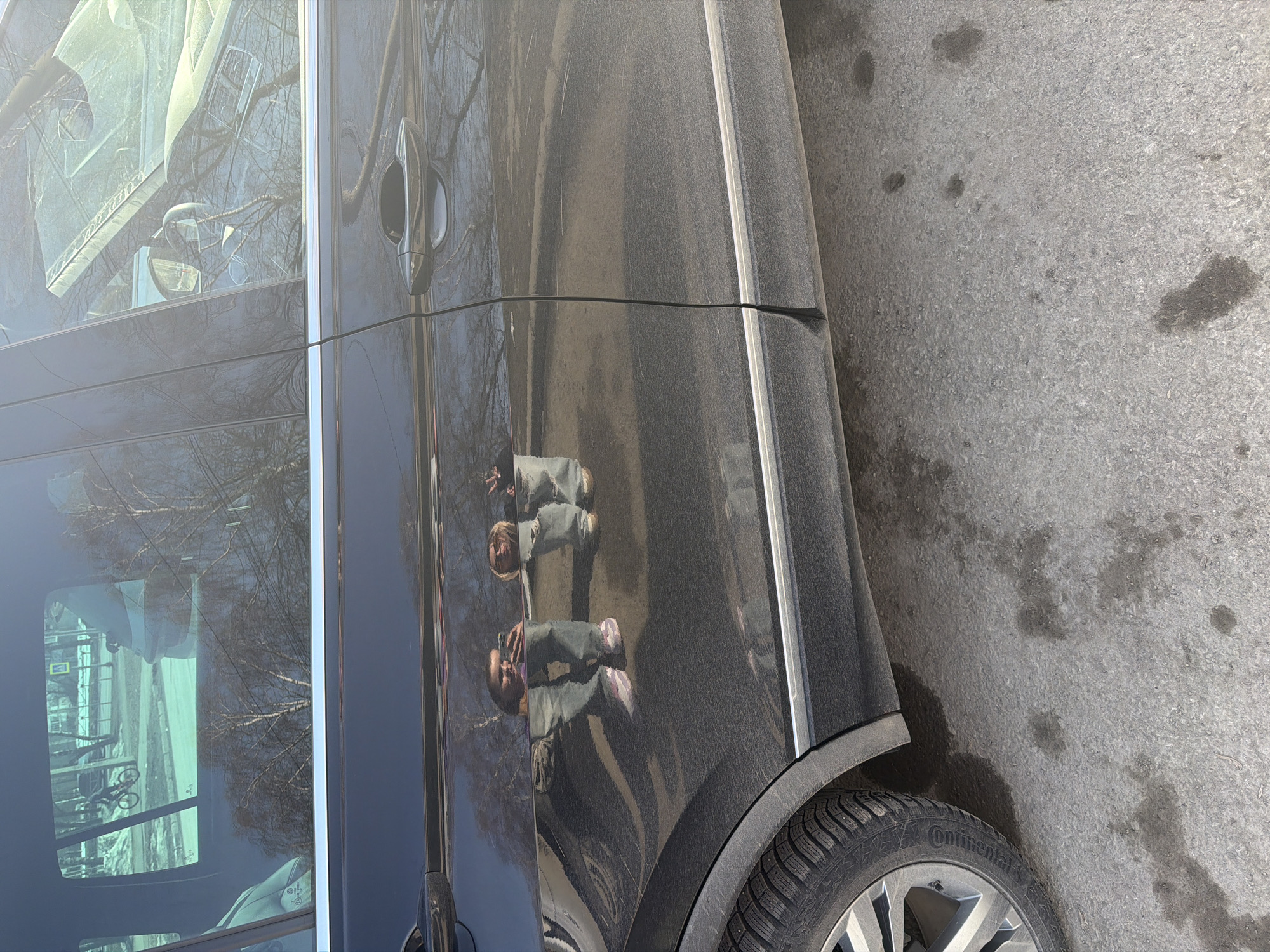 A close-up, slightly angled, outdoor shot depicts a black SUV parked on an asphalt surface. The vehicle's side is visible, including the driver's side door, a portion of the windshield, and the front tire. Reflections on the car's surface suggest an environment with trees and buildings. Reflected figures of two individuals, one shorter than the other, are visible on the car door. The tire has the word "Continental" visible on it. The background asphalt is gray and mottled, showing signs of wear and possibly water marks. The windshield reflection includes street signs and a bicycle. The photo was taken in Berezniki, Russia.