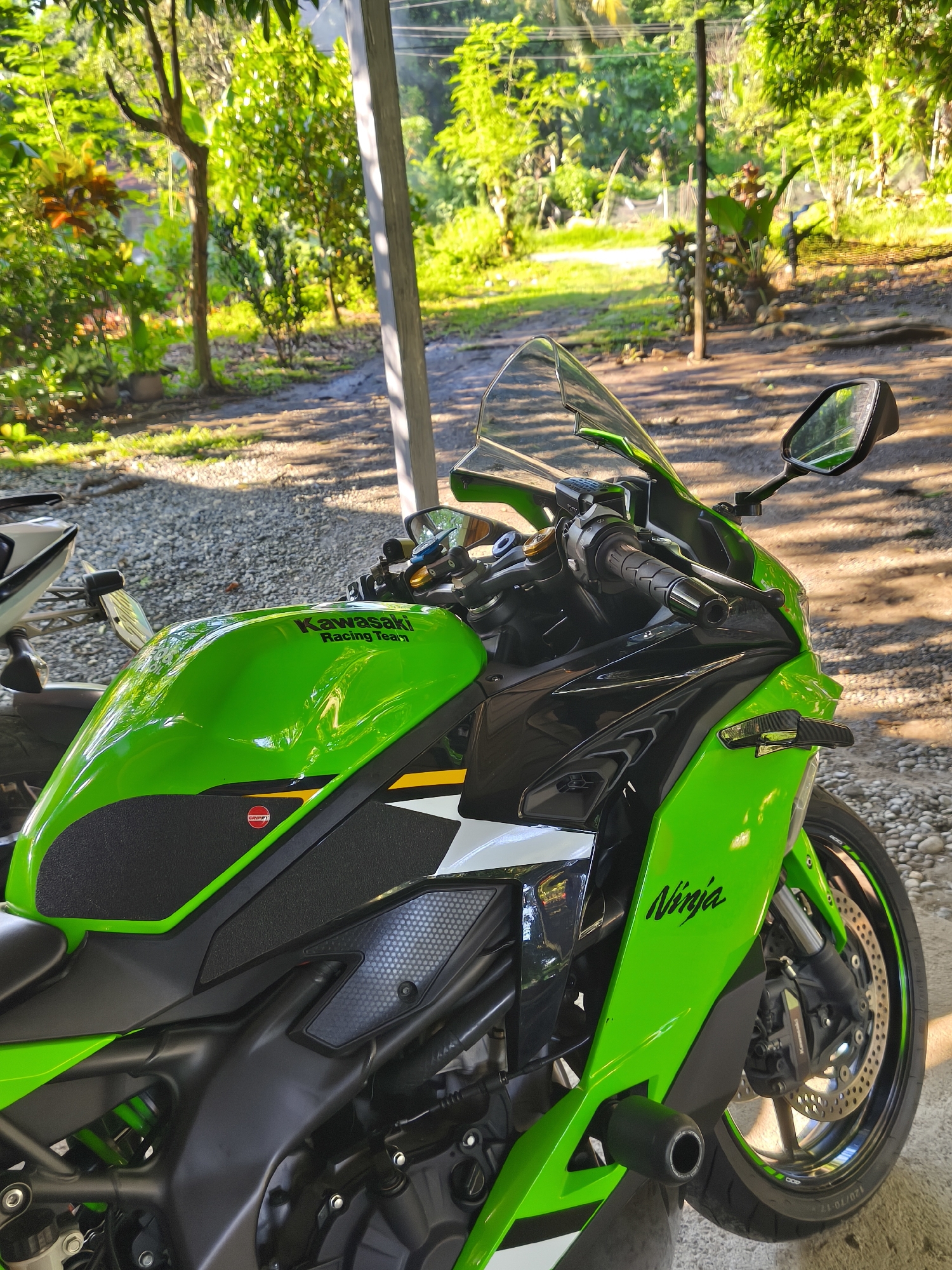 A vibrant green Kawasaki Ninja motorcycle is the central subject of the image. It is parked on a gravel surface, likely a driveway or parking area, with a partial view of another motorcycle to the left. The motorcycle is brightly lit, revealing details like the Kawasaki Racing Team and Ninja logos, as well as black tank grips. The background includes lush greenery, trees, and a glimpse of what appears to be a residential area or a park. The scene suggests an outdoor setting on a bright day. The location is not specified.