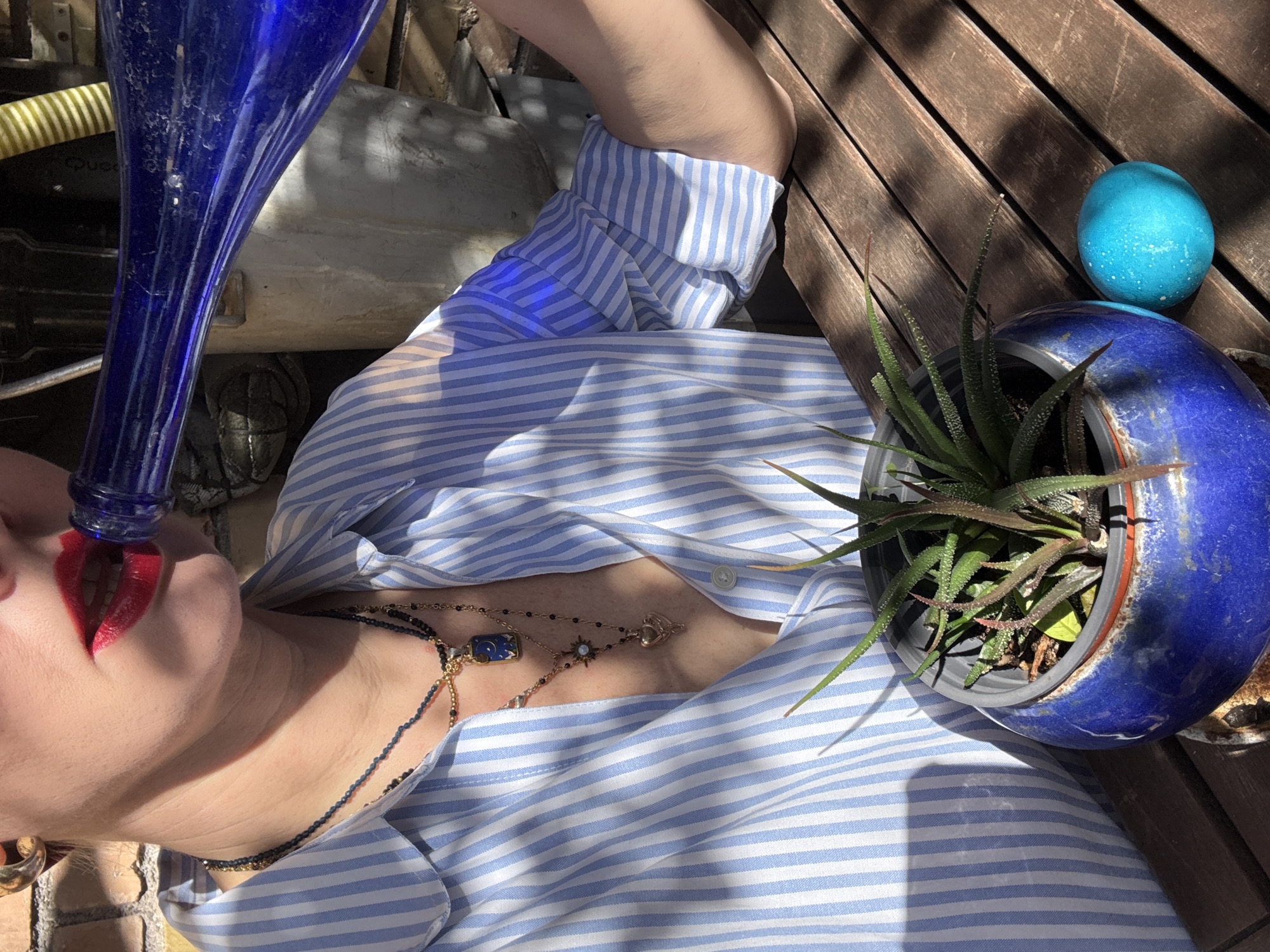 A top-down shot shows a person in a light blue and white striped shirt lying on a wooden surface, likely a deck. The person is holding a tall, dark blue glass bottle to their red-lipsticked mouth. The person is wearing several necklaces, including a gold chain and a beaded chain. A blue-glazed flowerpot containing a succulent-type plant is visible to the right of the person. Next to the flowerpot is a small, bright blue egg. The background features a yellow hose and other miscellaneous objects. There is no verifiable evidence that this image was taken in Rome, Italy.