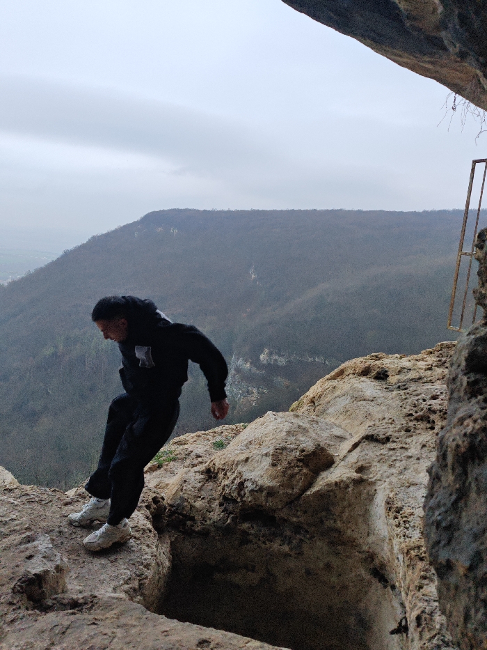 The image depicts a lone individual on a rugged, light-brown rocky cliff edge, overlooking a vast, forested valley under a uniformly overcast sky. The location is consistent with the Shumen Plateau, near the city of Shumen, Bulgaria.
The subject is a male, dressed in a black long-sleeved hooded top with a light-colored emblem on the left chest, black trousers, and light-colored athletic shoes. He is captured mid-action, leaning forward and slightly to his left, with his right foot planted on an elevated rock surface. His left leg is extended forward, positioned directly over a deep, dark recess or crevice in the rock immediately below him, indicating an active step or jump across an obstruction.
The immediate foreground consists of textured, uneven rock formations. To the far right, a partial view of a rusted, skeletal metal frame is visible, appearing to be anchored into the cliff face. The background presents multiple layers of hills and mountains covered in dense, dark green and brown vegetation, receding into the atmospheric haze under the grey sky. A small portion of a distant, possibly urban or agricultural area is faintly visible in the far left background.