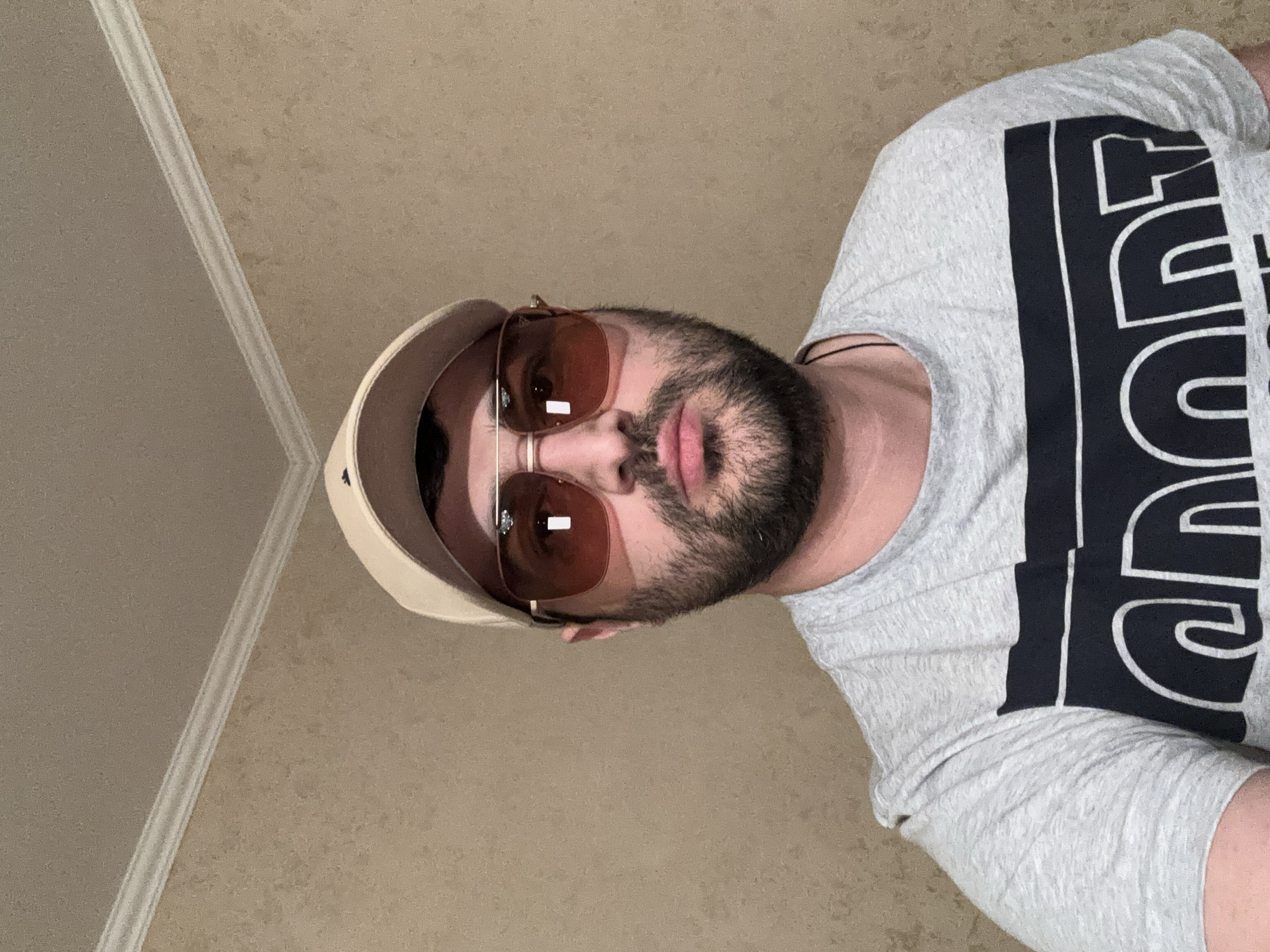 The image is a close-up, vertically oriented portrait of a man wearing a light-colored baseball cap backwards, brown-tinted aviator sunglasses, and a grey t-shirt with a large black graphic on the front. The man has dark hair and a beard, and his skin tone is medium. His eyes are not visible behind the sunglasses. His lips are slightly parted. He appears to be looking directly at the camera.
The background is a textured, light brown wall, with a white decorative molding or trim running diagonally from the top left corner of the image. The lighting suggests it is daytime, and the environment appears to be indoors, possibly a home. There is no clear indication of the weather or a specific location. The man's expression is neutral, and the overall mood of the image is casual.
The black graphic on the t-shirt consists of bold, outlined letters. While the full text is not entirely visible, parts of the letters "SPORT" can be discerned, with the letters "S", "P", "O", and "R" clearly readable, and possibly the end of a "T". There is also a thin black cord visible around the man's neck, possibly a necklace.