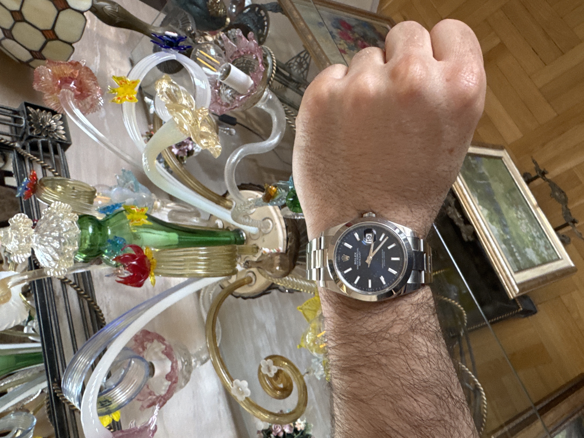 A man's forearm and hand are positioned in the center of the frame, with a stainless steel wristwatch on his wrist. The watch has a black dial, silver case, and a silver bracelet. The background is a cluttered glass table with various decorative objects, including a Tiffany-style lamp and several glass ornaments. A framed picture leans against the glass table, and a parquet floor is visible in the background. The location is likely indoors, but there is no additional information available to infer it is in Plovdiv, Bulgaria. The forearm is noticeably hairy. The man's fist is clenched. The lighting appears to be ambient.