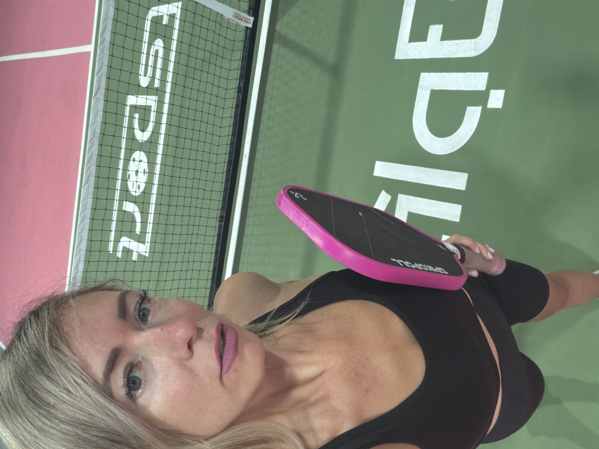 A woman with blonde hair and blue eyes is taking a selfie on a pickleball court. She is wearing a black sports bra and black shorts, and holding a pink pickleball paddle. The court is green with white lines, and there is a net in the background. The word "ESPORT" is visible on the net. The court appears to be indoors, given the lighting and the netting. This scene does not appear to be in Da Lat, Vietnam, as there are no specific visual cues to suggest this location.