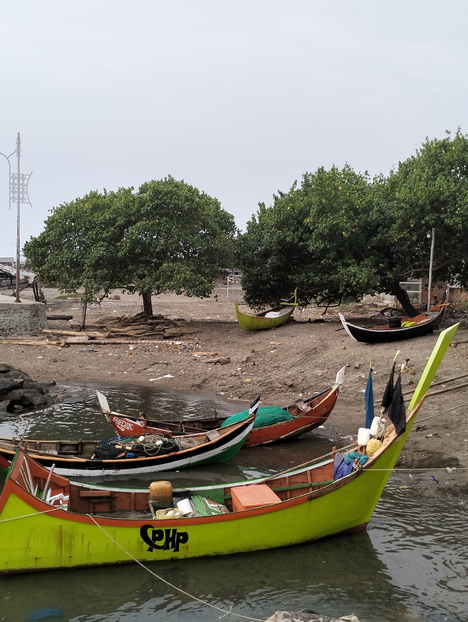 The image depicts a waterfront scene in Lhokseumawe, Indonesia, under an overcast sky. The foreground features dark, rippled water where multiple traditional wooden fishing boats are moored. The most prominent boat, in the lower left, is predominantly lime green with an orange upper trim and black lettering "PHP" on its side. It contains a yellow jerrycan and other unidentified objects. Behind it, a red and black boat, partially submerged, holds a green fishing net. Further back, another red boat with "SIEDAK" visible on its stern is also in the water.
The shoreline consists of a light brown, sandy, and muddy beach heavily littered with debris, including pieces of wood, plastic, and general trash. Several large, dense green trees with exposed roots dominate the midground of the shoreline. Two other boats are beached on the shore: one is partially visible in light green on the left, and another, black with a white trim, rests under the trees on the right.
In the far background, indistinct structures, potentially shacks or small buildings, are visible on the left, along with a tall, decorative streetlamp pole. To the right, a red-brick structure is partially obscured by trees. No discernible human figures are present in the scene.