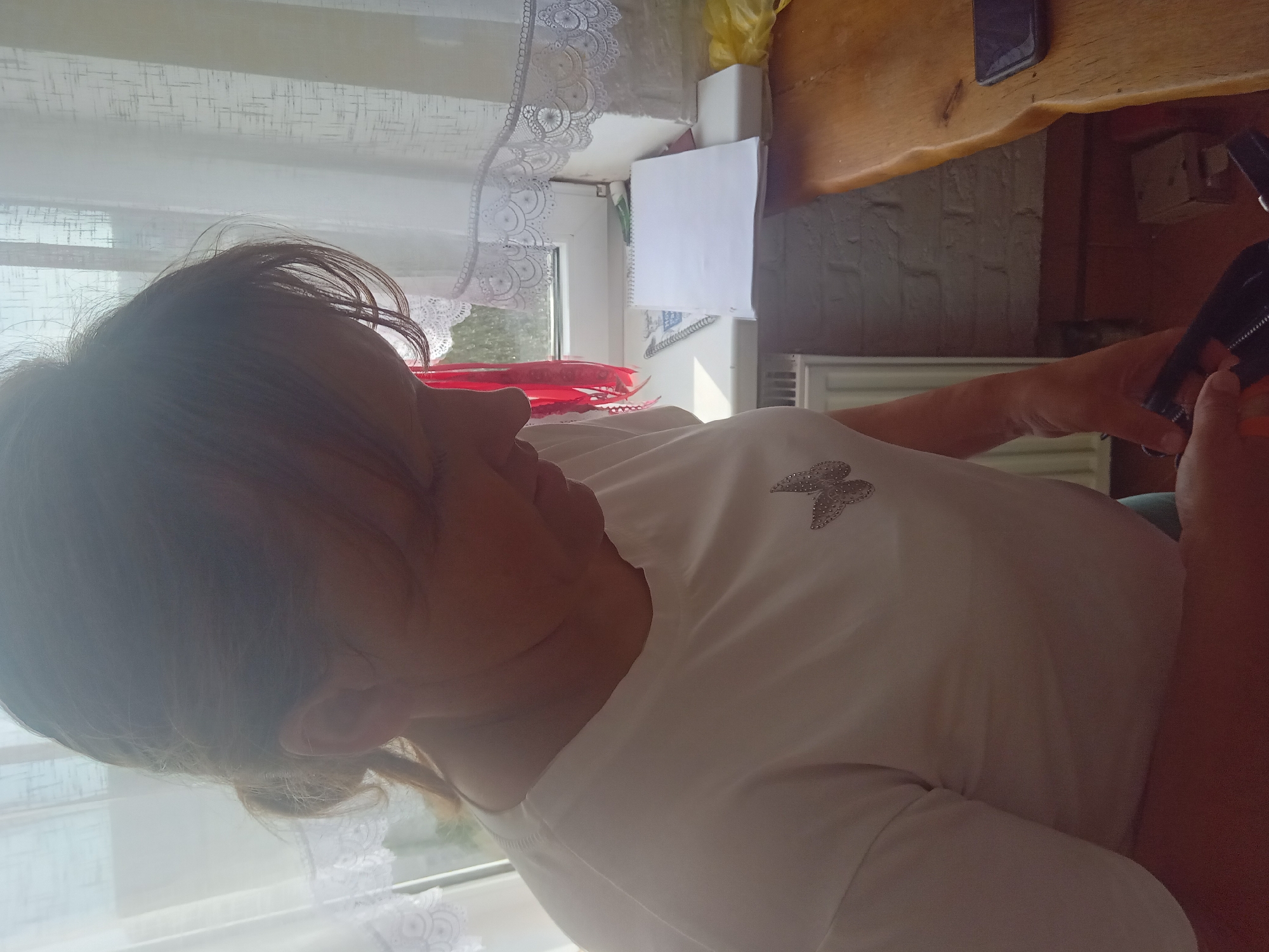 A woman with brown hair tied back is looking down and to her right. She is wearing a white t-shirt with a silver butterfly emblem on the left side of her chest. She is standing in front of a window with white curtains. To her right, on a wooden surface, are a spiral notebook, a yellow plastic bag, and a smartphone. The environment appears to be indoors, possibly a residential setting, given the window, curtains, and radiator visible in the background.