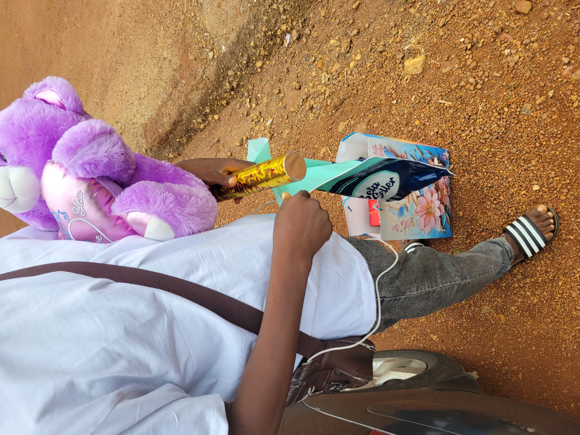 In Durumi, Nigeria, a person wearing a white t-shirt and grey jeans stands on a dirt ground. They are holding a large purple teddy bear with a pink heart that says "Love You." In their right hand, they also hold a gold cylindrical object and a light blue sheet of fabric. The fabric is partially tucked into a colorful gift bag with floral designs. A brown crossbody bag is slung over their left shoulder. The person is wearing black and white striped sandals. Part of a car is visible to the left of the frame. The ground is covered in reddish-brown dirt and small pebbles.