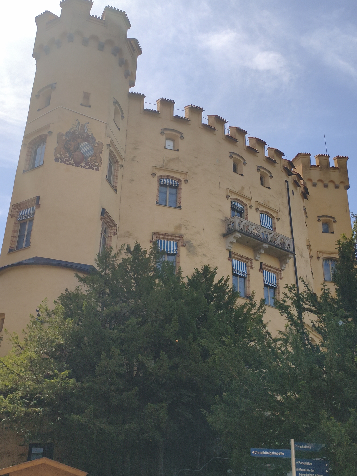 The image shows a low-angle view of Hohenschwangau Castle in Schwangau, Germany, bathed in sunlight under a partly cloudy sky. The castle's pale yellow facade dominates the frame, with its distinctive crenellated battlements and a prominent tower on the left. A large coat of arms is painted on the tower. Several windows with blue and white striped awnings are visible along the castle walls, and a decorative balcony juts out from the upper floor. Lush green trees partially obscure the lower portion of the castle. In the lower right corner, a signpost with German text points to "Parkplätze," "Christkönigskapelle," and "Museum der bayerischen Könige," indicating parking and nearby attractions.