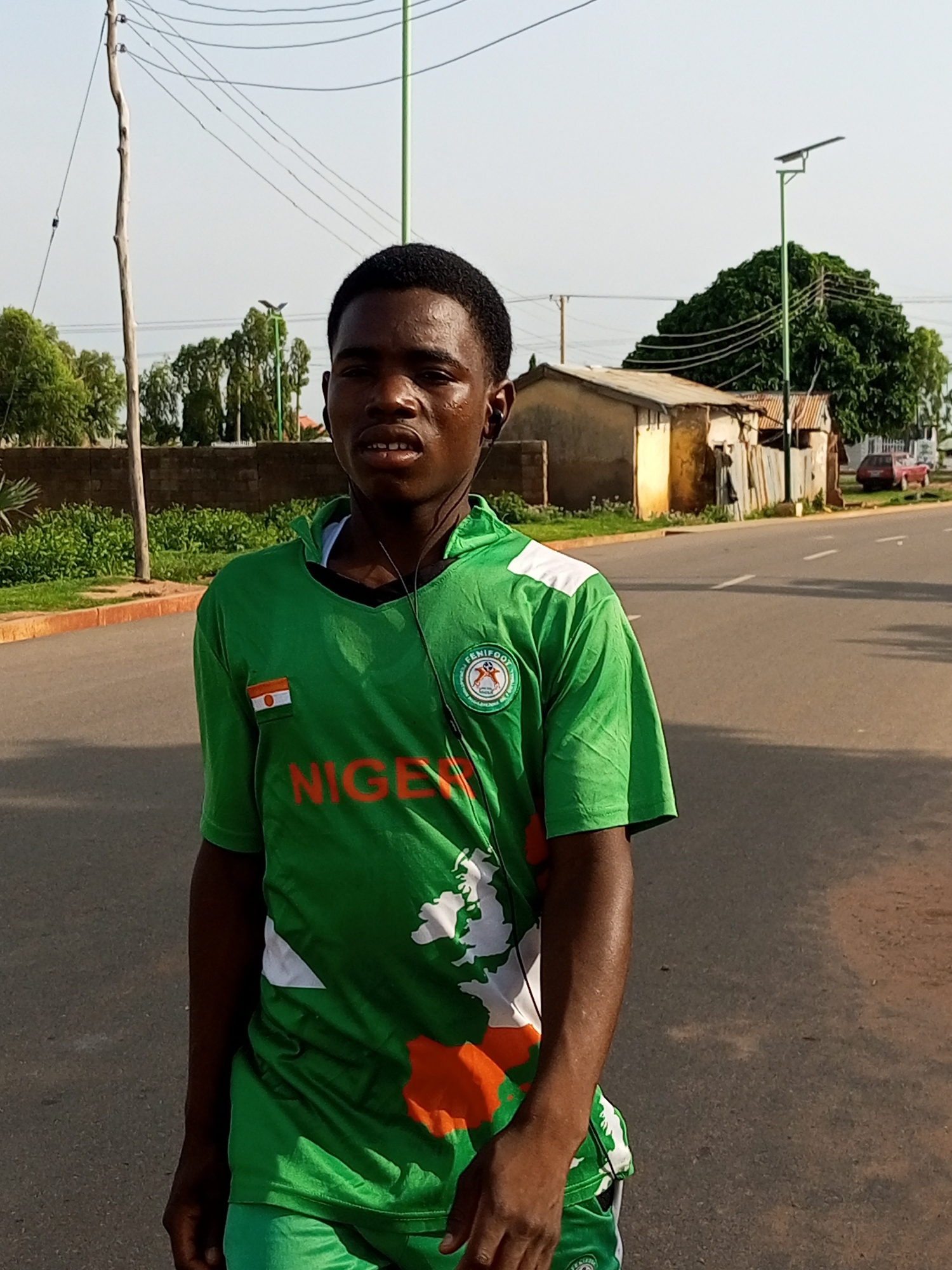 A young Black male, wearing a green Nigeria football jersey with white accents and an orange emblem, stands facing forward on a paved road in Tumfure, Nigeria. The jersey has "NIGERIA" emblazoned in orange letters across the chest, with a map graphic of the country also depicted. A small Nigerian flag is visible on the left chest of the jersey. He is wearing dark headphones that are plugged into his ears, with the cord running down his front. His expression is neutral, with his mouth slightly open. The road appears to be a two-lane street with white dividing lines. To the left of the road, there is a short, orange-topped concrete barrier, followed by lush green vegetation and a brick wall. In the background, several buildings with corrugated metal roofs are visible, along with trees. A street light with a solar panel is positioned on the right side of the road. The sky is clear and bright, suggesting daytime. A red car is parked in the background on the right. A wooden utility pole with electrical wires is on the left side of the frame.