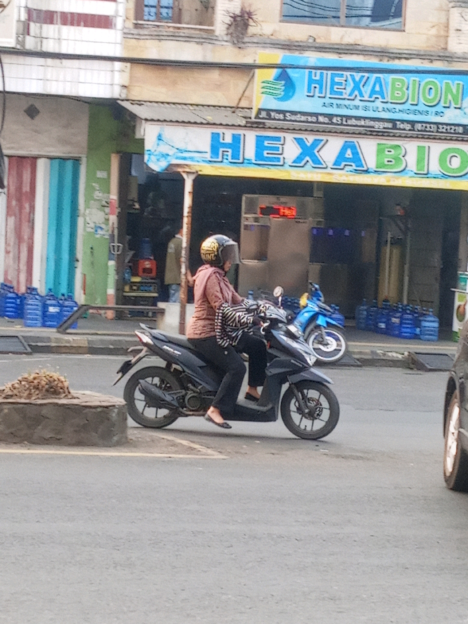 A person wearing a helmet and a patterned shirt is riding a black scooter down a paved street. The person's legs are visible, and they are wearing black pants and black shoes. The scooter has a license plate that is unreadable. In the background, there is a building with a sign that reads "HEXABION" in large blue and green letters. Below this sign, there is another sign that says "AIR MINUM ISI ULANG HIGIENIS / RO". The building also has several large blue water jugs stacked outside. To the left of the building, there are two motorcycles, one blue and one black, parked side-by-side. There is also a gray car visible on the right side of the image. The street appears to be in a commercial area, with shops and businesses lining the road. The overall scene suggests a typical day in a bustling Indonesian city.