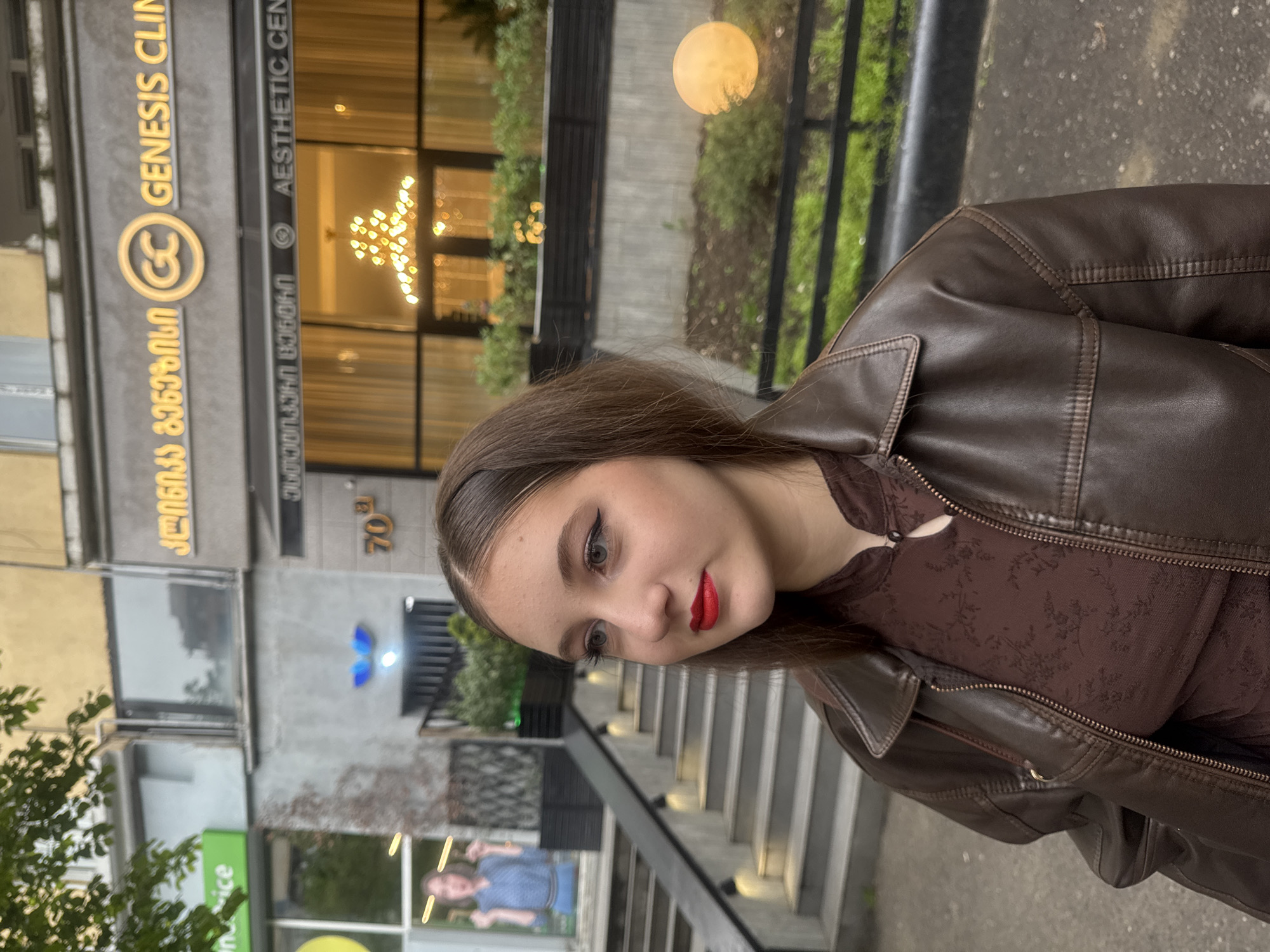 A young woman with brown hair and red lipstick stands outdoors. She wears a brown leather jacket over a darker brown floral-patterned top. Behind her, a building displays a sign with Georgian script alongside the English words "GENESIS CLINIC" and "AESTHETIC CENTER." The building address, "70a," is also visible. A staircase and partial views of other businesses are in the background. The scene suggests an urban environment, specifically Tbilisi, Georgia.