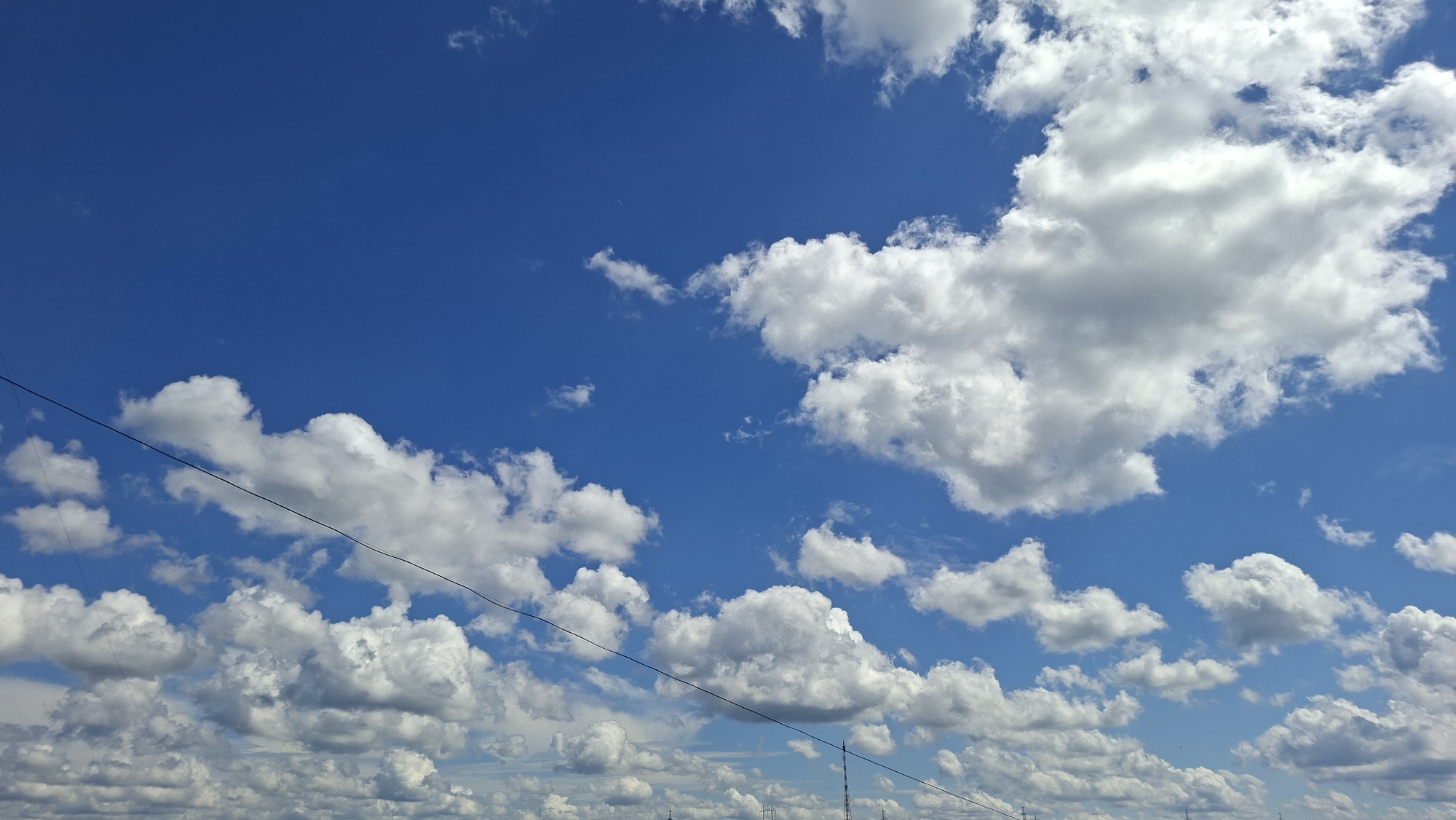 This media file depicts a bright, clear blue sky dotted with fluffy white cumulus clouds. The sun is shining, suggesting it is daytime. The clouds are scattered across the sky, with some appearing larger and more defined than others. There are thin, dark lines traversing the sky, which appear to be power lines or telephone wires. These wires suggest a connection to human civilization, possibly indicating a location near a town or rural area. The bottom of the frame shows a sliver of a horizon with some faint structures that could be buildings or towers, further reinforcing the idea of a populated area. The overall impression is one of a pleasant, sunny day with a picturesque sky. The location context of Chaikovsky, Russia, places this scene in a specific geographical area. There is no visible text in the image.