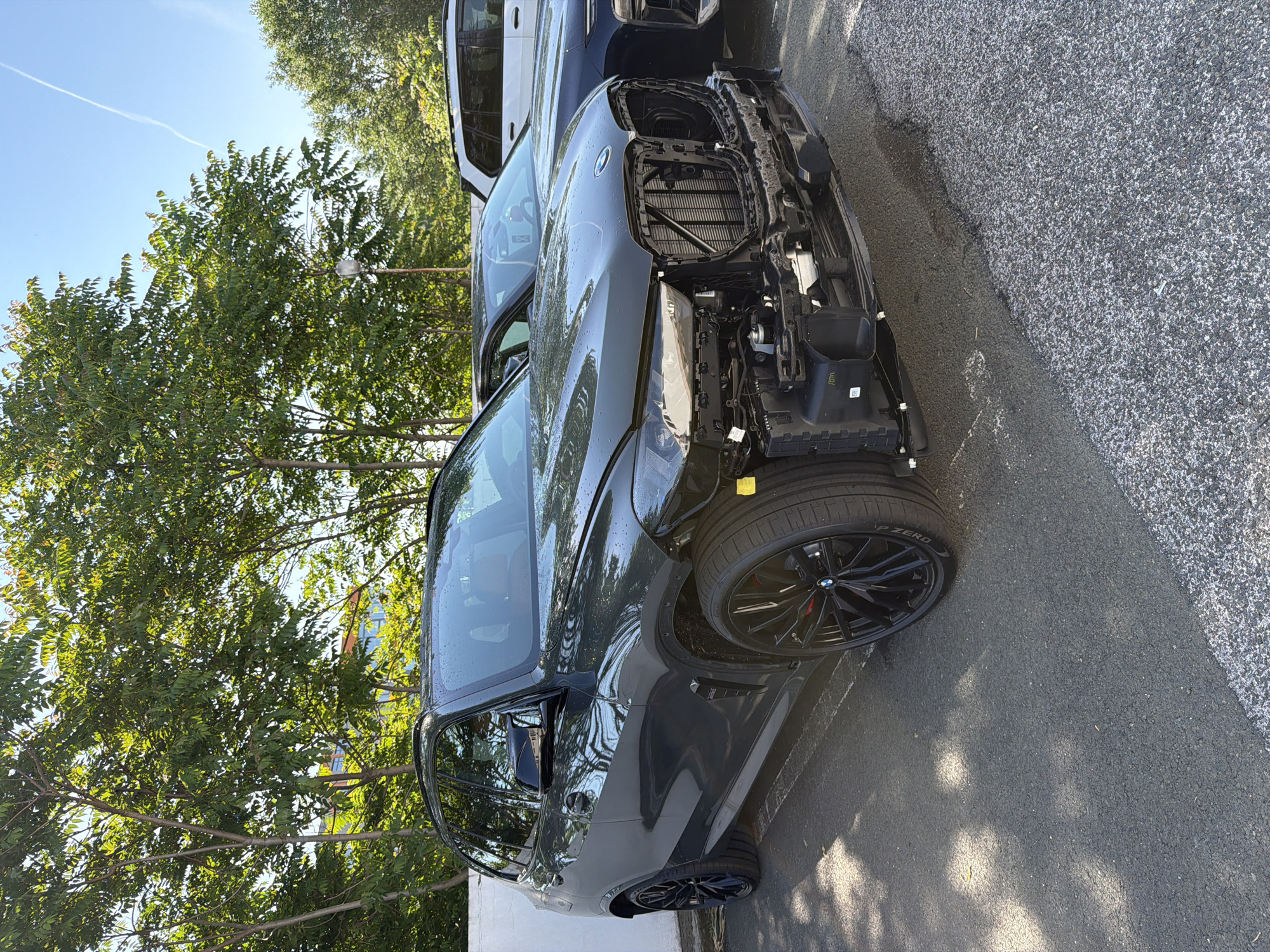 A dark grey SUV, likely a BMW, is parked outdoors on an asphalt surface, missing its entire front bumper. The vehicle's front structure, radiator, and part of the passenger-side headlight assembly are exposed. The driver's side front wheel is visible, featuring a black multi-spoke rim, red brake caliper, and a Pirelli P Zero tire. Water droplets are visible on the car's bodywork, suggesting recent rain or washing.
Behind the dark grey SUV, a white vehicle, possibly another SUV, is partially visible. The setting is bright and sunny, indicated by a clear blue sky with a single white contrail and distinct shadows. Lush green trees dominate the background, with a street light fixture noticeable among their branches. The asphalt ground has varying textures, appearing darker and smoother closer to the vehicle and lighter and coarser further away. A small white object, possibly a screw, lies on the lighter asphalt.
Visible text includes "P ZERO" on the tire sidewalls, a BMW logo on the wheel hubcaps, and alphanumeric codes like "372M" on a yellow sticker near the front wheel arch and "017M" on a white sticker on an internal component near the front right wheel.
The scene depicts a vehicle in a state of repair or damage, parked in an outdoor, likely urban or suburban environment in Burgas, Bulgaria, during daytime.