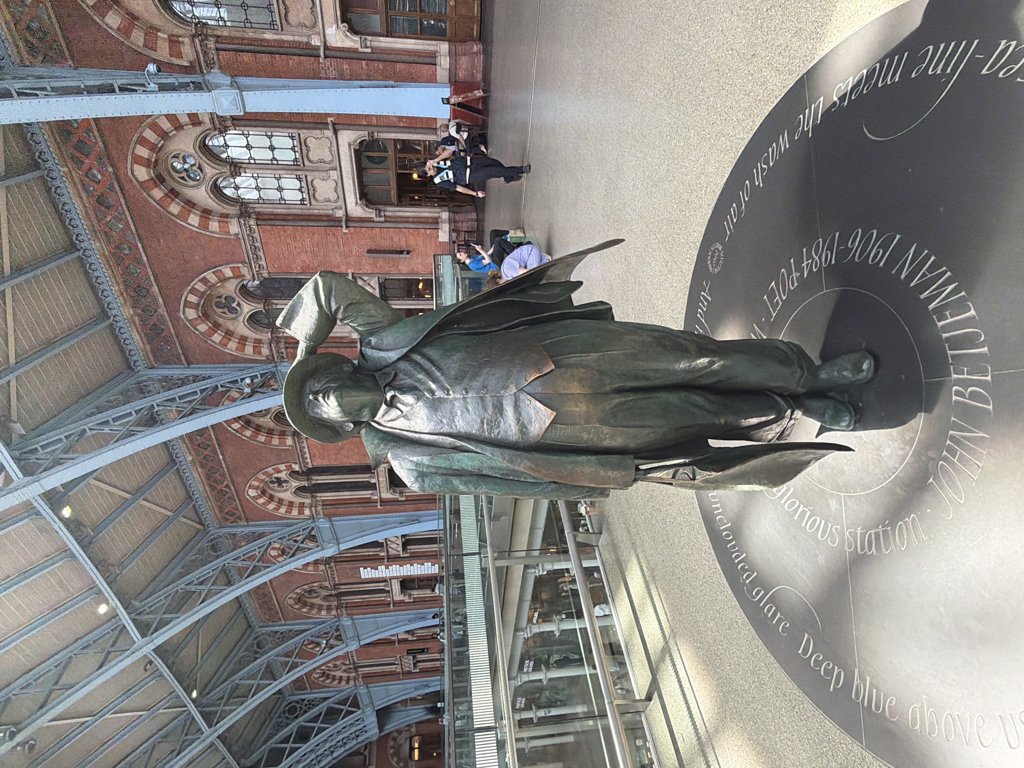 Here's a detailed description of the image:
The image is taken inside St Pancras International train station in London, United Kingdom. The focus is on a bronze statue of John Betjeman, a British poet and writer. The statue is positioned on a circular platform with inscribed text around its edge: "JOHN BETJEMAN 1906-1984 POET. Where the sea-line meets the wash of air. And in the shadowless, unclouded glare Deep blue above us… glorious station.”
The perspective is such that the station's vaulted roof, with its intricate ironwork and brickwork, is visible in the background. There are people walking in the background, including uniformed officers. There are glass barriers in the background. The lighting in the station appears bright.