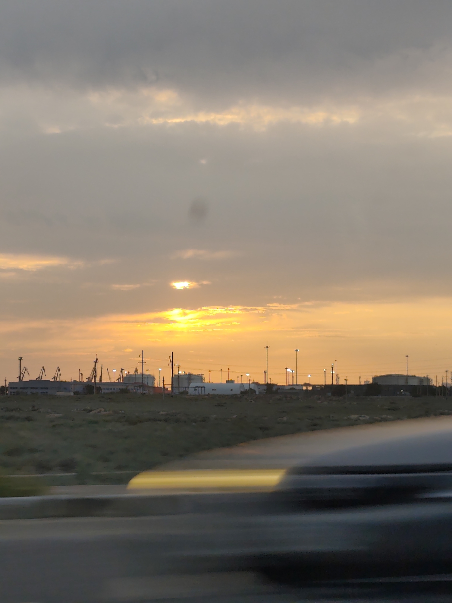 The image shows a sunset over an industrial area in the city of Ömirzaq, Kazakhstan. The sky is filled with a mix of gray clouds and bright orange and yellow hues from the setting sun. Several tall cranes are visible in the distance, along with industrial buildings and storage tanks. The foreground is blurred due to motion, suggesting the photo was taken from a moving vehicle. A portion of a vehicle is visible in the bottom of the frame, also blurred. The overall scene depicts a twilight industrial landscape under a dramatic sky.