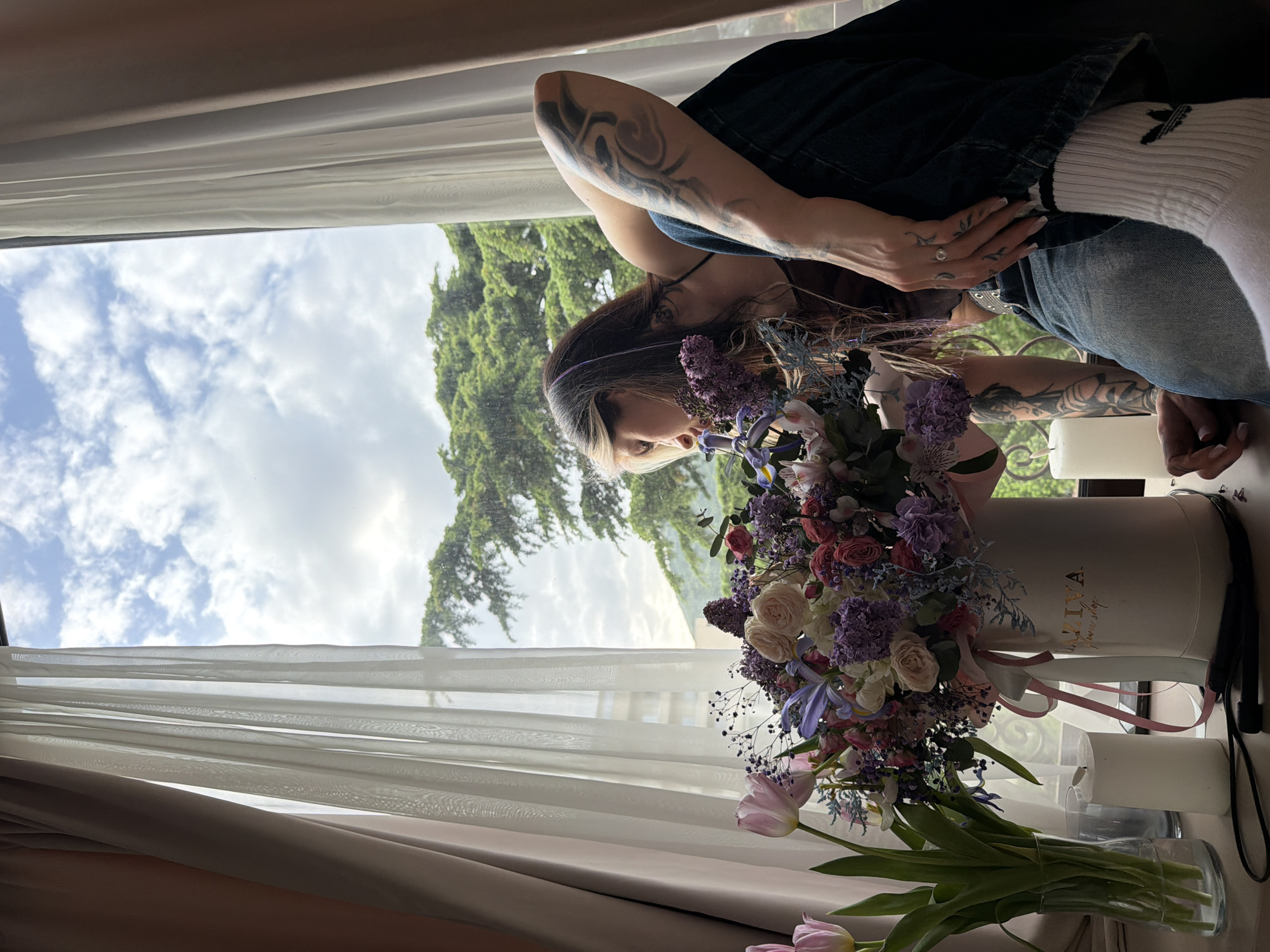 The image portrays a serene indoor scene by a large window, centered around a young woman admiring a vibrant floral arrangement. The woman, positioned to the right, has long dark hair with lighter streaks and is wearing a dark denim outfit. Her arms are notably covered with extensive tattoos featuring swirling, abstract designs. A ring is visible on her left ring finger, and her fingernails have a white polish. She looks down at the flowers with a gentle, appreciative expression.
The main focal point is a lush, colorful bouquet displayed in a white, round hatbox. Faint gold text, "AZIVA flower shop," is visible on the side of the hatbox. The bouquet consists of a rich variety of flowers including pink and white roses, purple hyacinths or lilacs, blue irises, and delicate baby's breath. In the foreground, several pink tulips are arranged separately in a clear glass vase. Two white pillar candles are also present on the surface next to the hatbox.
The setting is bright and airy due to a wide window on the left, draped with sheer white curtains. Through the window, a bright sky with scattered white clouds is visible, along with dense green trees, suggesting a natural, possibly hilly landscape characteristic of Budva, Montenegro. The scene is illuminated by natural daylight, indicating it is daytime.