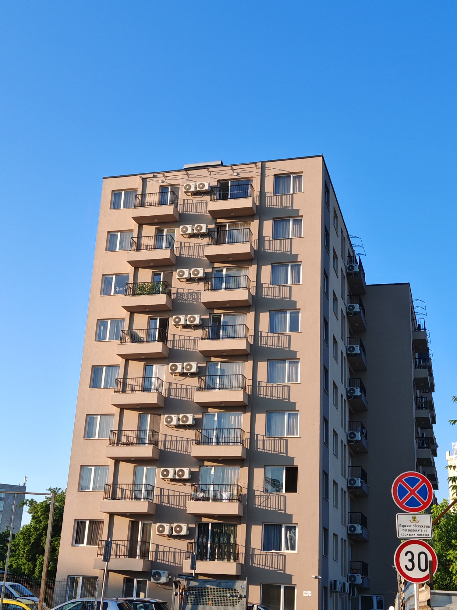 A tall, light brown, multi-story apartment building with many balconies and air conditioning units is the primary subject. The building has a modern architectural style with dark railings on the balconies and numerous windows. A clear blue sky dominates the upper portion of the image. In the foreground, there are traffic signs and parked cars, suggesting an urban environment. The traffic signs include a prohibition sign with a blue circle and red cross, a rectangular sign with text in Bulgarian that reads "ПАРКИНГ ОБСЛУЖВАЩ ПОЛЗВАТЕЛИТЕ НА СОЦИАЛНИ ЖИЛИЩА" (Parking for users of social housing), and a circular speed limit sign indicating "30". A tree is partially visible on the left side of the frame. The scene appears to be a street view in Plovdiv, Bulgaria, likely a residential area.