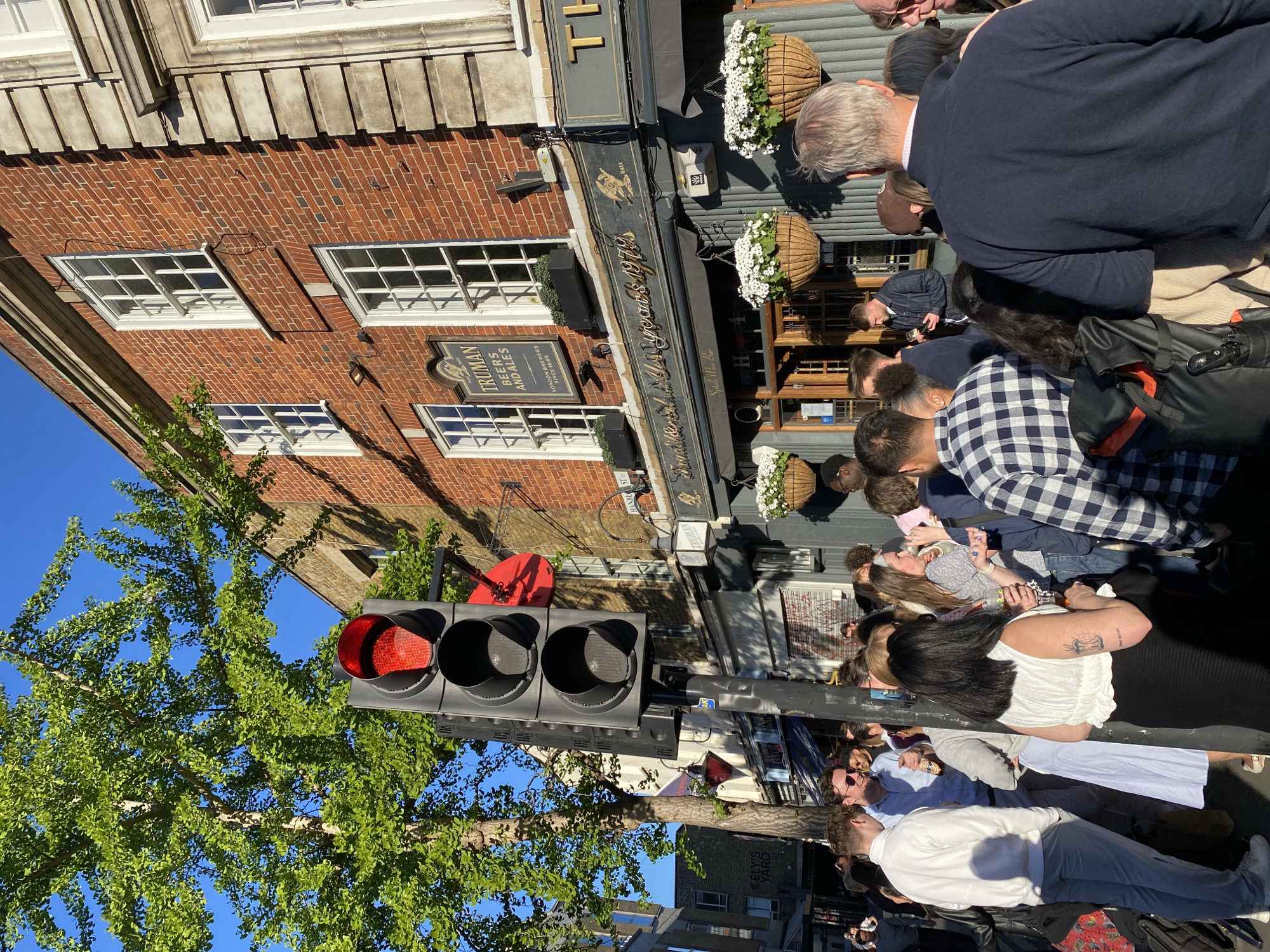 This outdoor street scene captures a vibrant gathering of people in London, United Kingdom, on a bright, sunny day. The focal point is a traditional red brick building housing a pub, identified by signs as "Spitalfields Wine Bar" and featuring "TRUMAN BEERS AND ALES LONDON BREWERS SINCE 1666." Numerous individuals, mostly adults, are casually standing and socializing on the sidewalk in front of the pub, with some facing towards the building.
The architecture includes classic sash windows with white frames set into the brickwork, and a decorative stone facade at the top of the building. The pub's entrance is adorned with hanging baskets filled with white flowers. To the left, a prominent traffic light displays a red signal, indicating a stop. A lush green tree with abundant leaves provides a natural element against the clear blue sky. In the background, another sign reads "ELY'S YARD," and a street sign partially shows "HANBURY ST." The strong sunlight casts distinct shadows, suggesting a mid-day to afternoon time. The overall atmosphere appears relaxed and social.