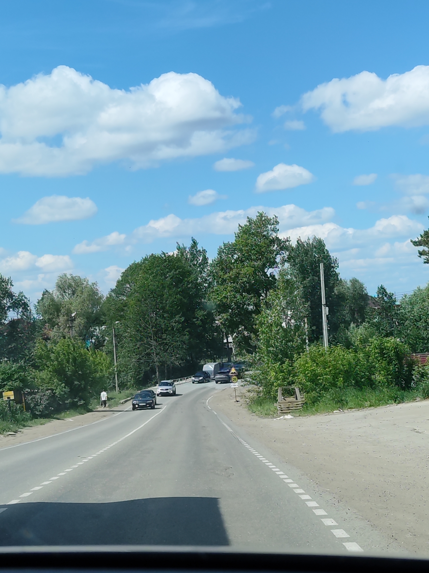 The image captures a daytime scene from inside a vehicle, looking ahead at a two-lane asphalt road winding through a green, semi-rural landscape under a bright blue sky. Numerous fluffy white cumulus clouds are scattered across the sky.
In the foreground, the dark dashboard or windshield frame of the vehicle is visible. The road features a dashed white line marking the center and a solid white line on its left edge. It gently curves to the right in the mid-distance.
Several vehicles are present on the road. A dark-colored sedan is in the left lane in the mid-ground, followed by a lighter-colored SUV. Further down the road, near the curve, a truck with a trailer and other cars are visible, suggesting traffic.
On the left side of the road, a person dressed in a light shirt and dark pants walks away from the viewer on a dirt shoulder. To their left, lush green trees and dense bushes line the road. A small yellow sign, partially obscured, is visible on the far left. A clearer view reveals a yellow sign with "20" and an upward arrow, likely indicating a 20 km/h speed limit or advisory.
The right side of the road is also lined with various green trees and bushes, with a dirt shoulder. Utility poles are visible interspersed among the trees on both sides. The overall impression is a sunny day on a road in a verdant, possibly rural or suburban, area of Chaikovsky, Russia.