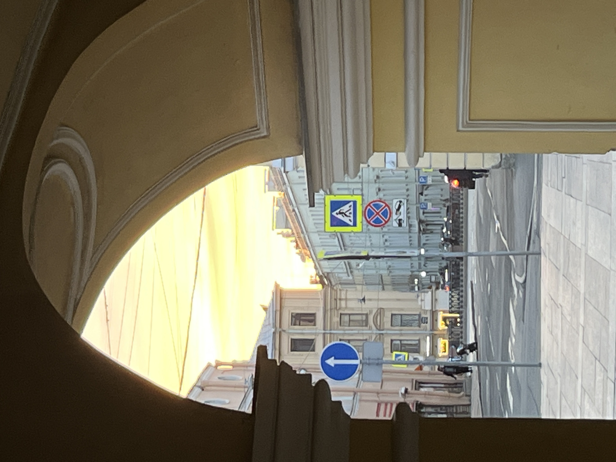 The image captures a street scene viewed through a wide, ornate archway or vaulted passage. The interior of the archway, dominating the foreground and left side, is a warm, muted yellow-ochre color, featuring decorative moldings.
Beyond the arch, a city street is visible under a strong, bright, yellowish light, suggesting either sunrise or sunset. Multi-story buildings with classical architectural features like cornices and structured windows line the street.
Several road signs are prominent. A square yellow sign with a blue border and a black pedestrian crossing symbol is visible near the center. Next to it is a circular red sign with a blue cross and a diagonal red line, indicating "No Stopping" or "No Standing." Further down, a circular blue sign with a white arrow pointing left signifies a mandatory direction. A traffic light, showing a red signal, is also seen. The street appears paved, with faint road markings. In the distance, two dark, indistinct figures, likely people, are visible on the street. The overall scene depicts a typical urban environment during the golden hour, likely in a European city given the architectural style and sign conventions. No legible text is present on the signs, only symbols.